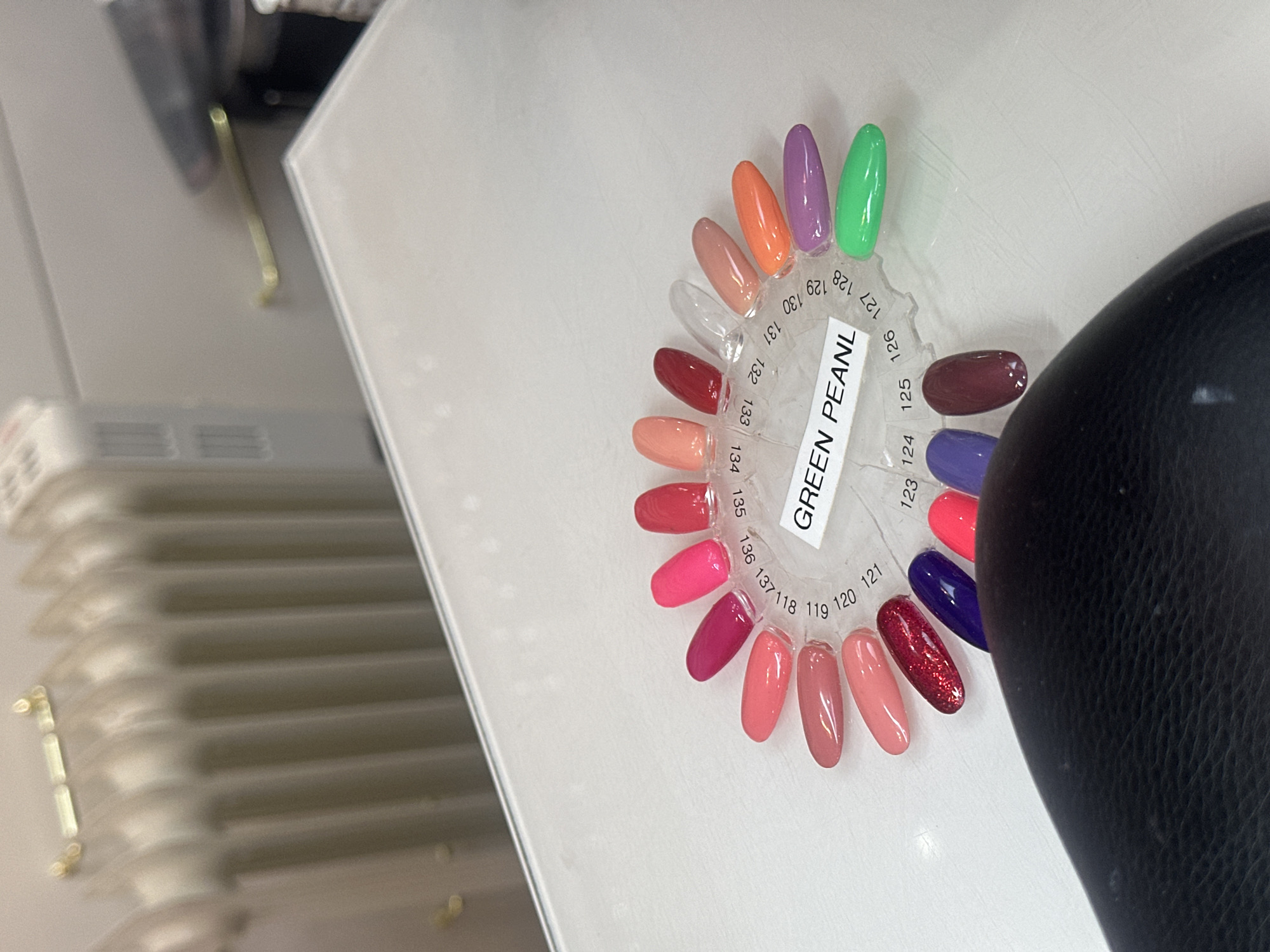 The image depicts a nail polish color sample wheel resting on a white surface, likely a table or counter. The wheel presents a variety of shades ranging from pinks and reds to purples, greens, and oranges. Each color is associated with a number, visible around the wheel's edge. A sticker in the center of the wheel reads "GREEN PEANL" (likely a misspelling). In the background, a blurred object possibly a chair or equipment is seen.
It is not possible to identify the location in Tunbridge Wells, United Kingdom from the visual information provided.