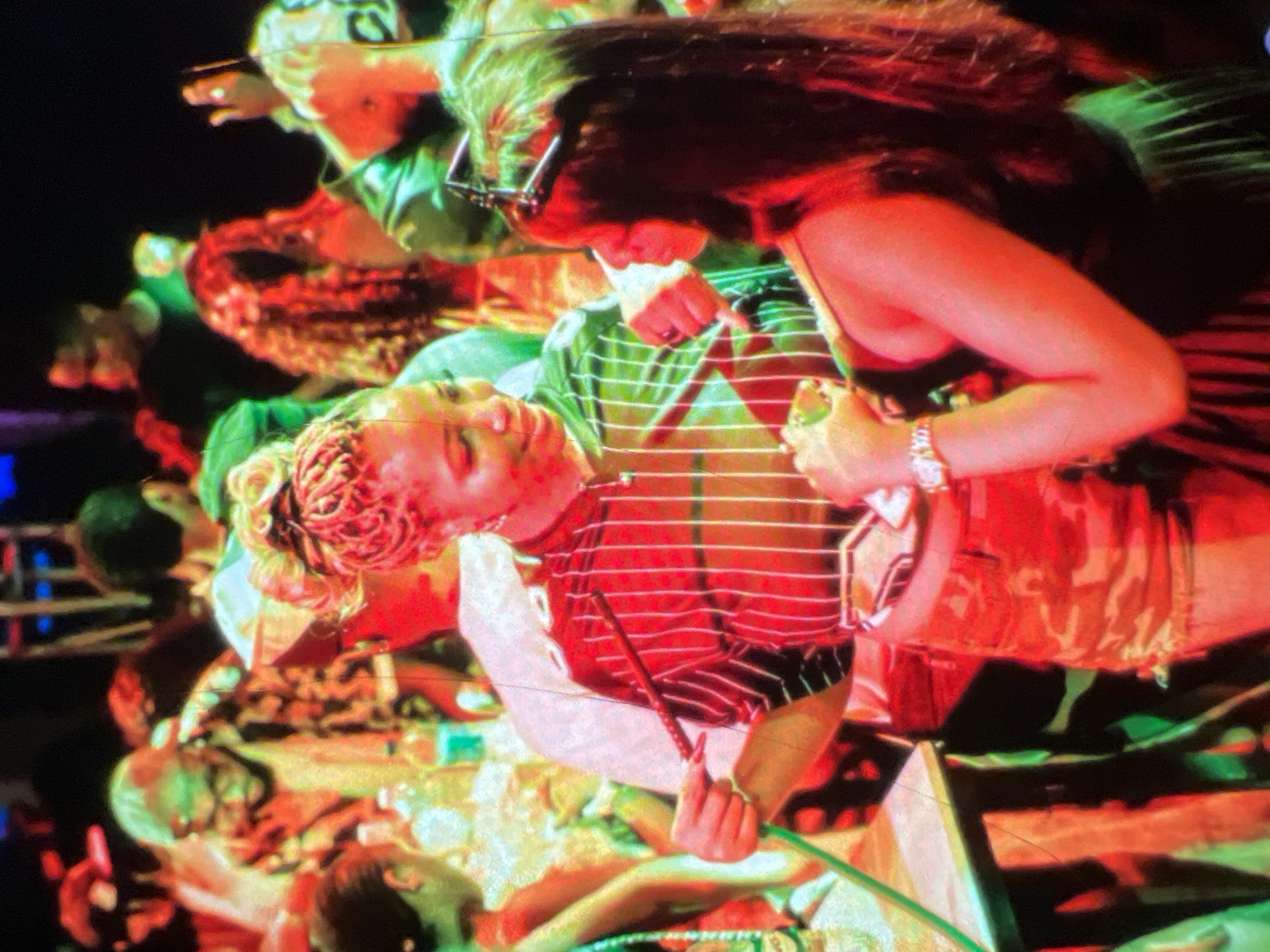 The image depicts a group of people at an indoor event, possibly a party or gathering, with vibrant lighting that casts reddish and greenish hues. In the foreground, two women are the central focus. The woman on the left, wearing a red and white striped shirt and camouflage shorts, is holding a dark slender object, possibly a wand or a decorative item, in her right hand, and a green hose-like object in her left. She has blonde hair styled in braids and is looking towards the woman on her right. The woman on the right, with long dark hair, is wearing a black dress and is looking at the woman on her left. Her left arm is visible, adorned with a watch. In the background, other individuals are present, some with patterned clothing and glasses, contributing to the crowded and lively atmosphere. The presence of bottles and what appears to be a table suggest a social gathering. The scene is highly stylized due to the lighting, creating an almost ethereal or dramatic effect. The mention of Lagos, Nigeria, in the context of this image suggests that the event is taking place in that city, though no specific landmarks or contextual clues within the image itself confirm this location.