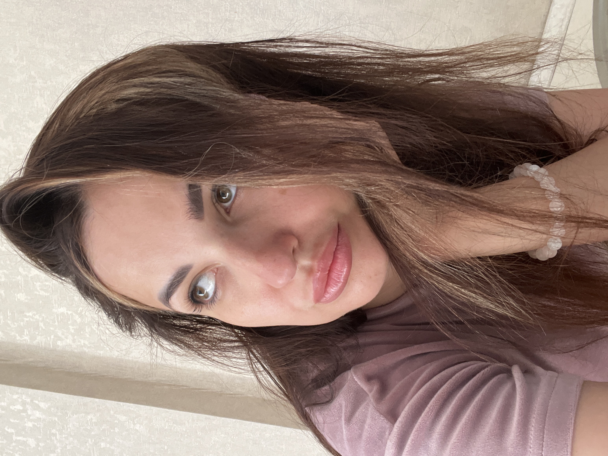 A woman with long, dark brown hair with lighter streaks is looking up and to her left, resting her head on her hand which is adorned with a beaded bracelet. She is wearing a soft, pink, long-sleeved shirt. The background is a textured, light-colored wall. The image is a close-up of her face and upper body.