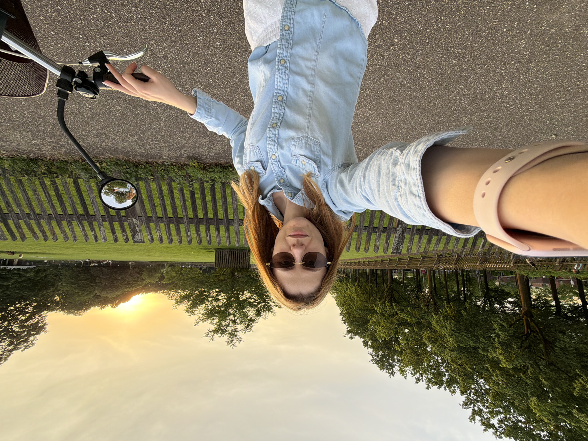 A fair-skinned person wearing sunglasses, a light blue denim shirt, and a white top, is positioned in the center of the image, holding the handlebars of a bicycle. The background displays a park-like scene with a wooden fence separating a green lawn from a paved path. The sun is setting, casting a warm glow over the landscape. The bicycle's mirror reflects the scene behind the person. The paved path suggests a road or walkway. The scene resembles an outdoor area possibly within the city of Helmond, Netherlands.