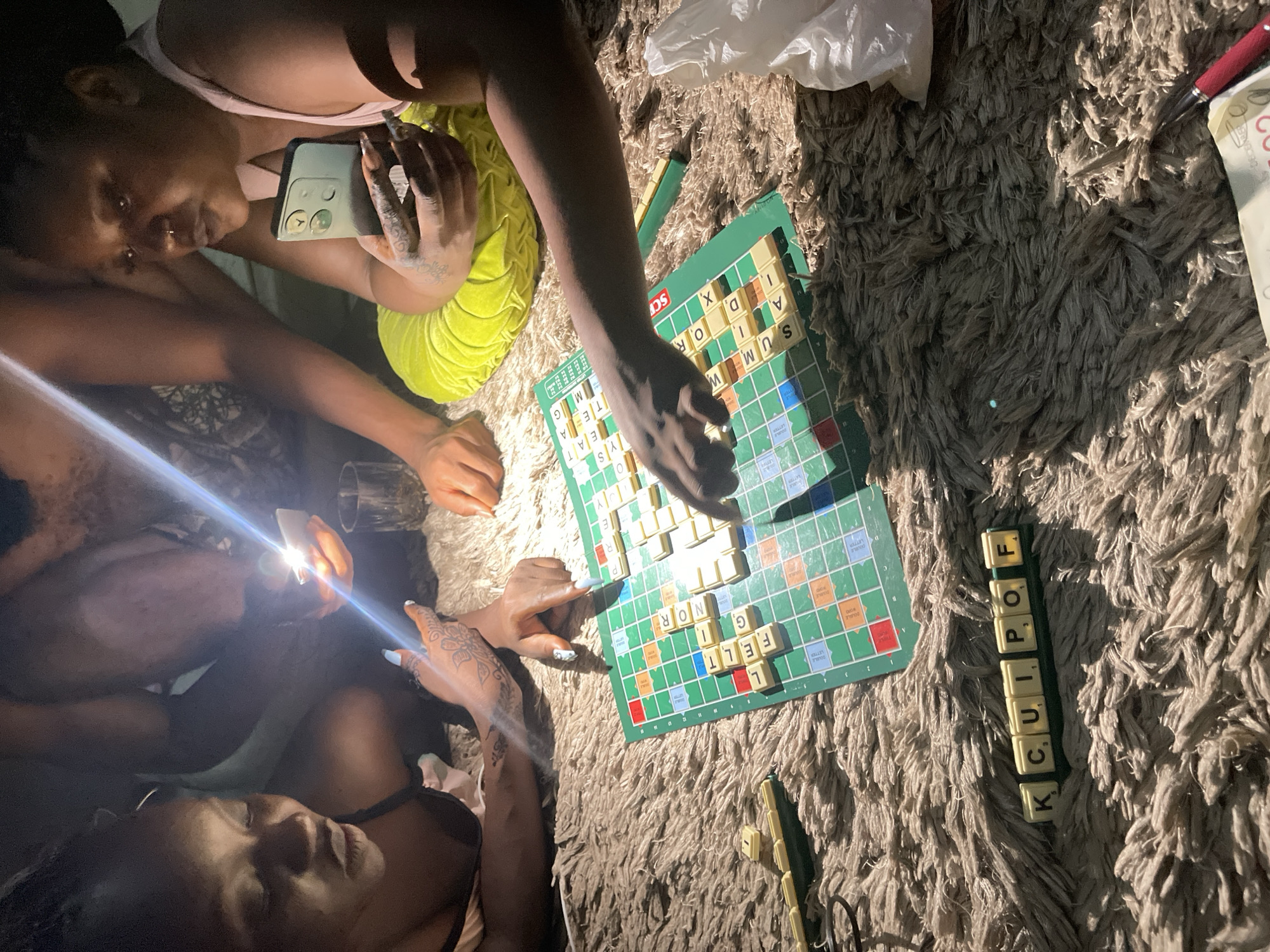 This media file captures an intimate, dimly lit indoor scene in Kuchingoro, Nigeria, where two individuals are playing Scrabble on a shaggy, light-brown rug. The overall impression is one of a casual evening or nighttime activity, necessitated by the prominent use of a smartphone flashlight for illumination.
The subjects include two people, likely women, engaged in the board game. One person, positioned in the upper frame, leans over the Scrabble board, holding a smartphone, and wears a light top with bright yellow-green attire. Their hand, adorned with intricate mehndi patterns and long nails, is reaching towards the board. The second person, lying in the lower foreground, holds a smartphone with its flashlight directed at the game, providing a strong beam of light across the scene.
The central object is a green Scrabble board with numerous letter tiles forming words such as "SUM," "OR," "TEA," "FELT," and "NOR." A tile rack adjacent to the board holds the letters "KCUIPOF." Other objects scattered on the rug include a white plastic bag, a red and silver pen, and a glass. The atmosphere appears relaxed and focused, typical of a board game session. Visible text from the OCR includes "SUM," "OR," "TEA," "FELT," "NOR," and "KCUIPOF" on the tiles, along with a partial "DECEMBER" on a piece of paper.