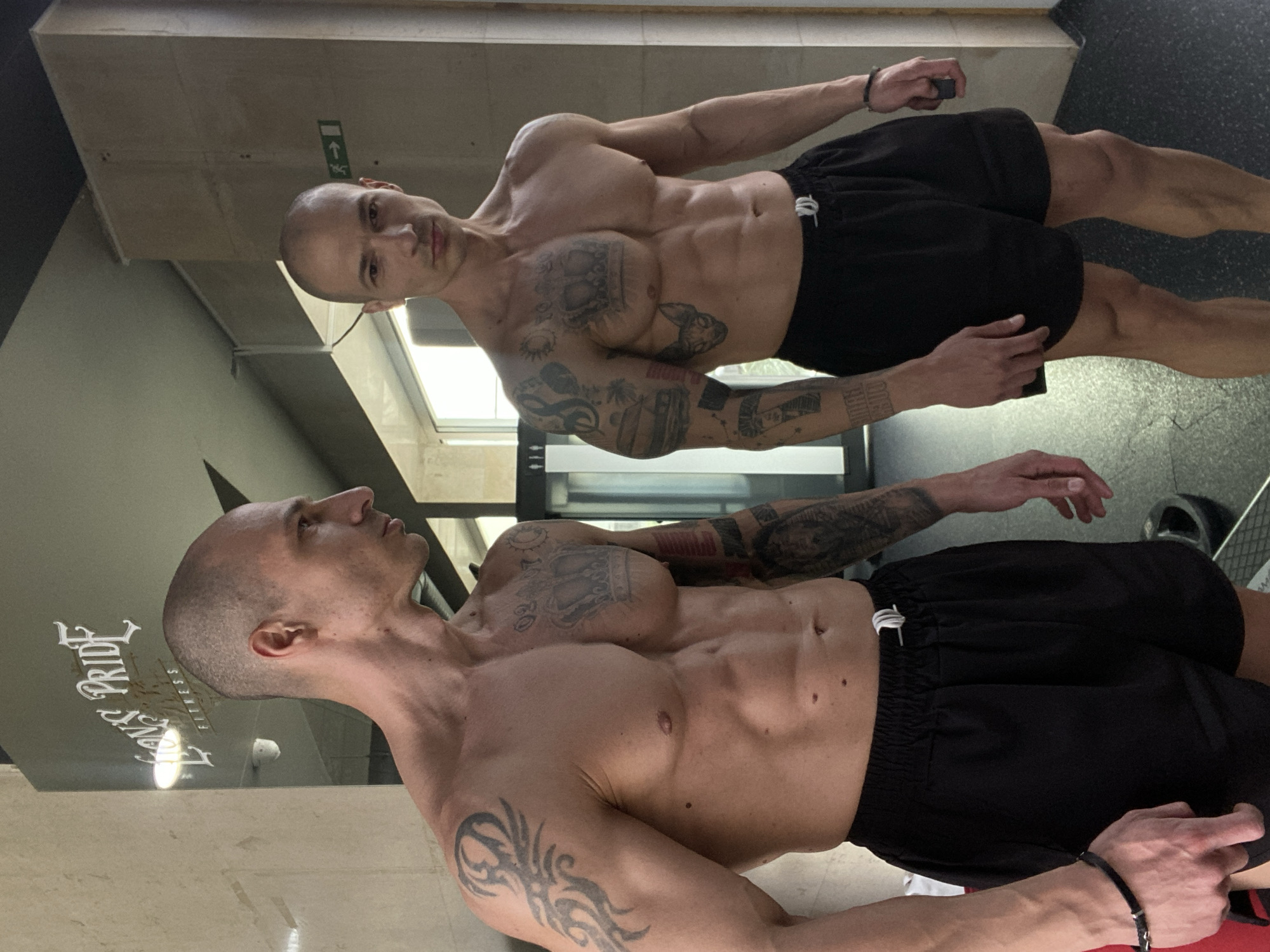 A muscular, bald man with tattoos on his upper body is standing in front of a reflective surface. He is wearing black shorts. His reflection is visible in the surface, creating a mirror image of him. The reflection shows the man from a slightly different angle. The background features a wall with text that includes "Pride Fitness." The location is likely a fitness center or gym in Pleven, Bulgaria.