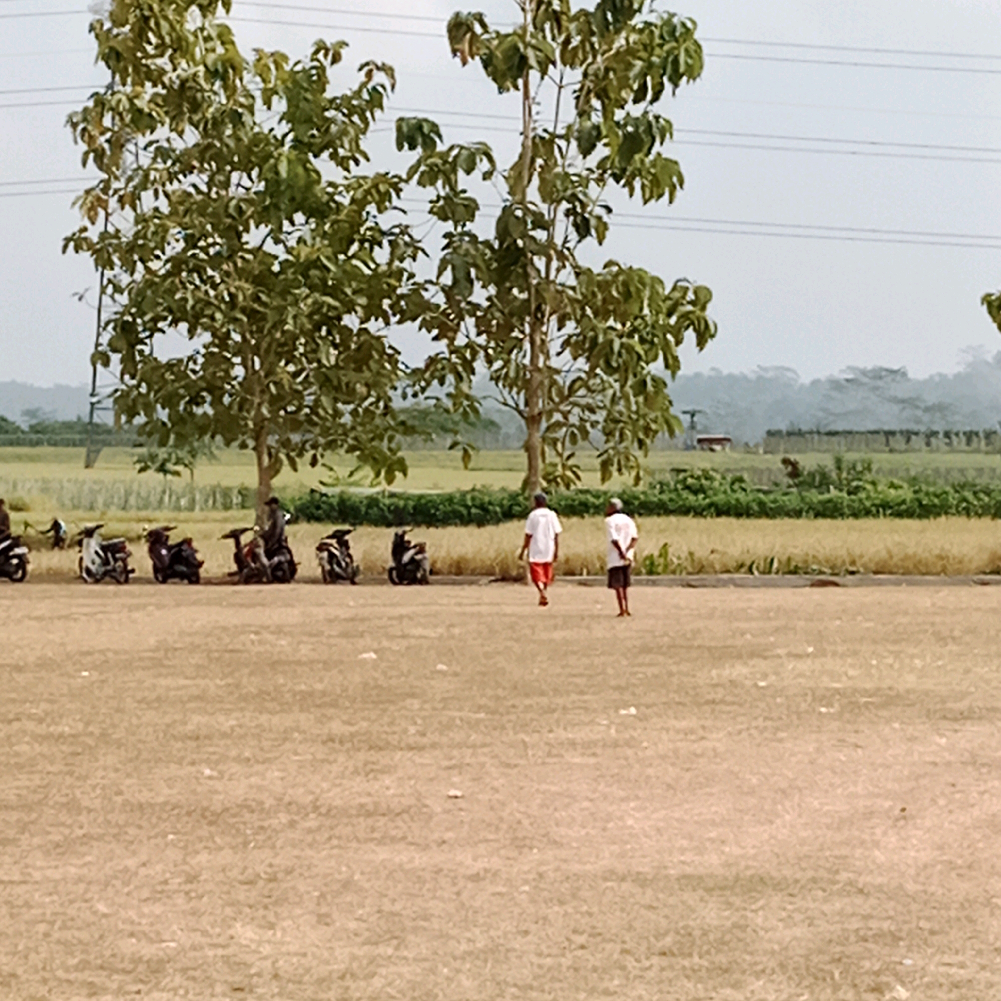 This is a photograph taken in the city of Rakit, Indonesia. The image depicts an outdoor scene with a dry, brown dirt field in the foreground. In the middle ground, a line of parked motorcycles is visible, with several individuals standing or walking nearby. Two men are the most prominent figures, both wearing white shirts. One is walking away from the camera, wearing red shorts, and the other is facing away, wearing dark shorts. Behind the motorcycles and people, a field of dry, golden-brown grass or crops extends into the background, bordered by a line of green bushes and trees. Further in the distance, a hazy landscape of hills and trees is visible under a pale sky. Power lines are also present in the upper portion of the image, running horizontally across the sky. The overall impression is of a rural or semi-rural environment in a dry season.