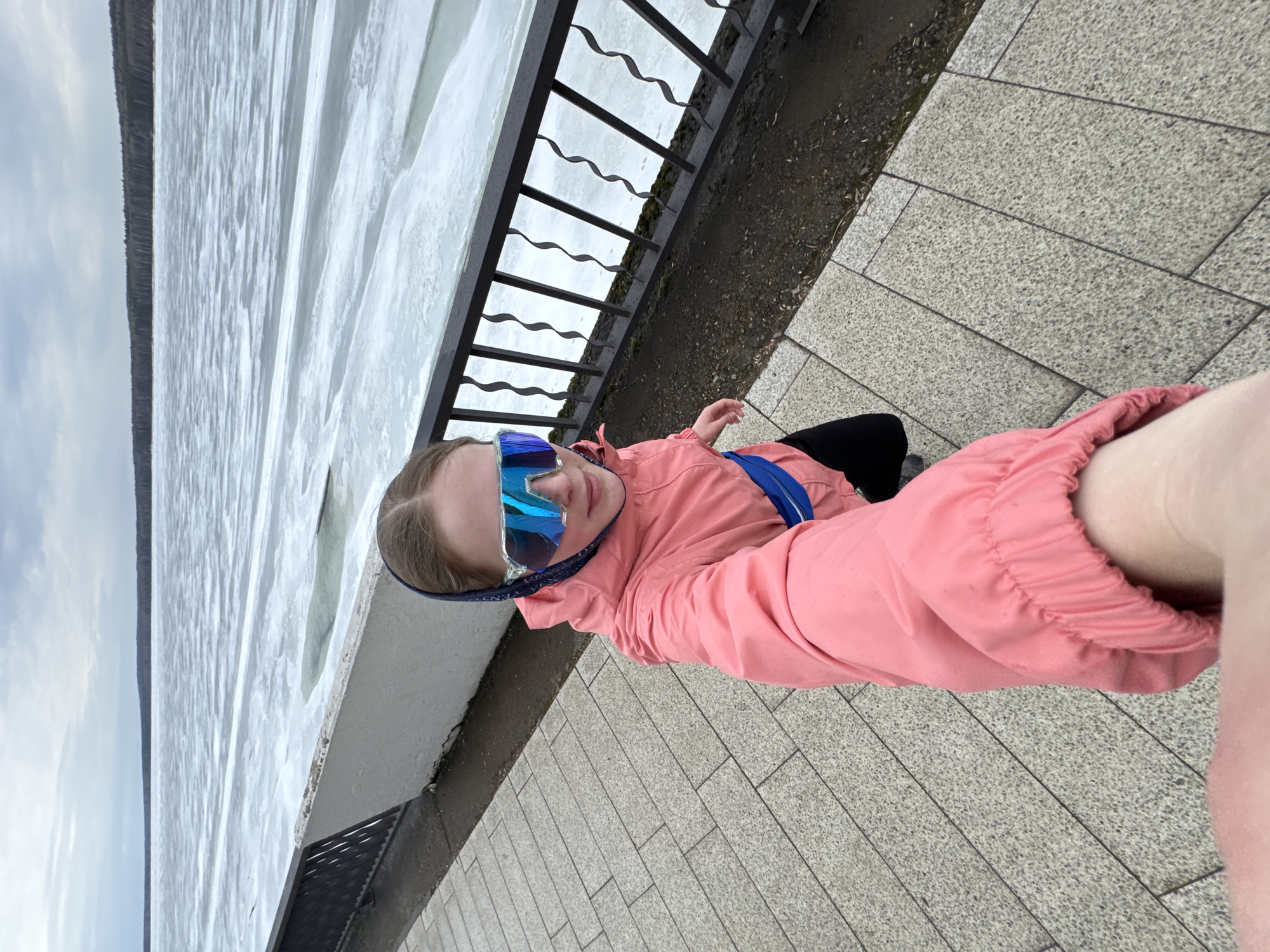 A young person with fair skin is seen in a tilted selfie, wearing a pink jacket and sunglasses. The background shows a body of water with a treeline on the horizon and a black metal railing. The setting is urban, indicated by the patterned paving stones on which the person stands. Without additional information or features characteristic to Chaikovsky, Russia, the specific location within the city cannot be determined.