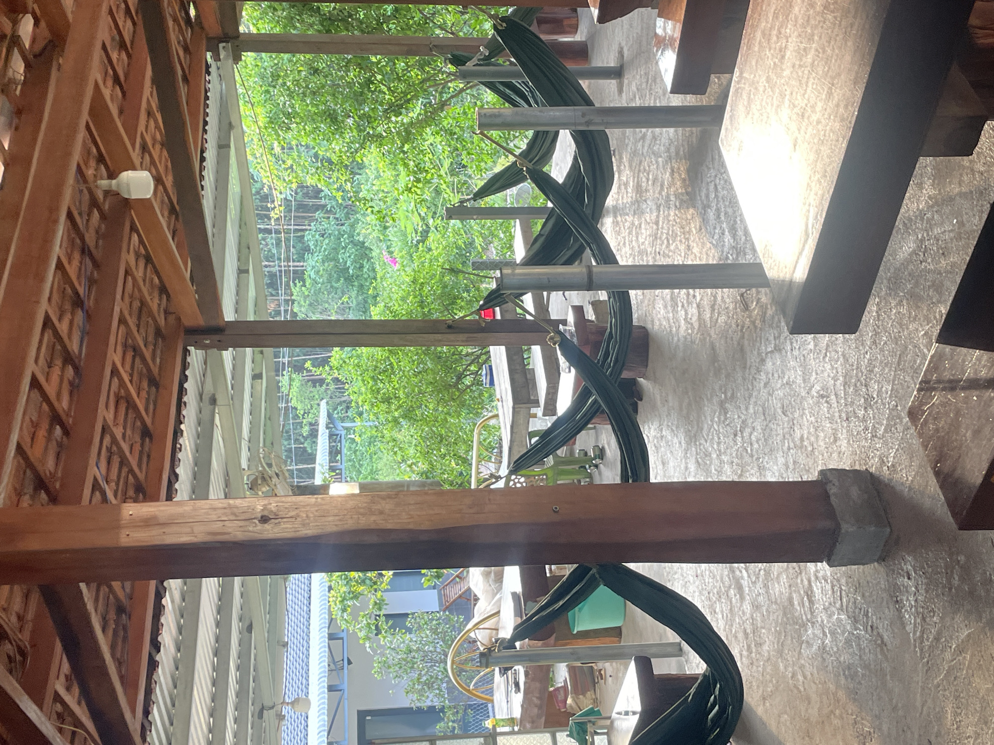 The image displays an open-air relaxation or dining area in Tây Ninh Hòa, Vietnam. The structure features a visible wooden rafter and terra cotta tiled roof layered above a corrugated metal roof. Multiple sturdy, square-section wooden columns, some with gray concrete bases, support the overhead structure. Several dark green hammocks are strung between these wooden columns and vertical gray metal poles; all hammocks appear empty.
The flooring consists of a textured, light-colored concrete surface, exhibiting both sunlit areas and shadows. Various rustic wooden tables and benches are arranged throughout the space. In the mid-ground, a large, lighter-colored table is visible, accompanied by a golden-framed chair and a light blue bucket. A green bottle is situated on one of the tables. The foreground includes a dark wooden table with a prominent reflection of sunlight. A single white, modern-style LED bulb hangs from the wooden roof structure.
Beyond the open sides of the facility, dense, vibrant green foliage and trees form the background, indicative of a lush natural environment. A faint pink flower is discernible within the distant vegetation. To the left background, a portion of a building with a light blue corrugated metal roof and white walls is visible. The scene is illuminated by natural sunlight, producing defined shadows and bright highlights across the surfaces. No individuals are present in the image.