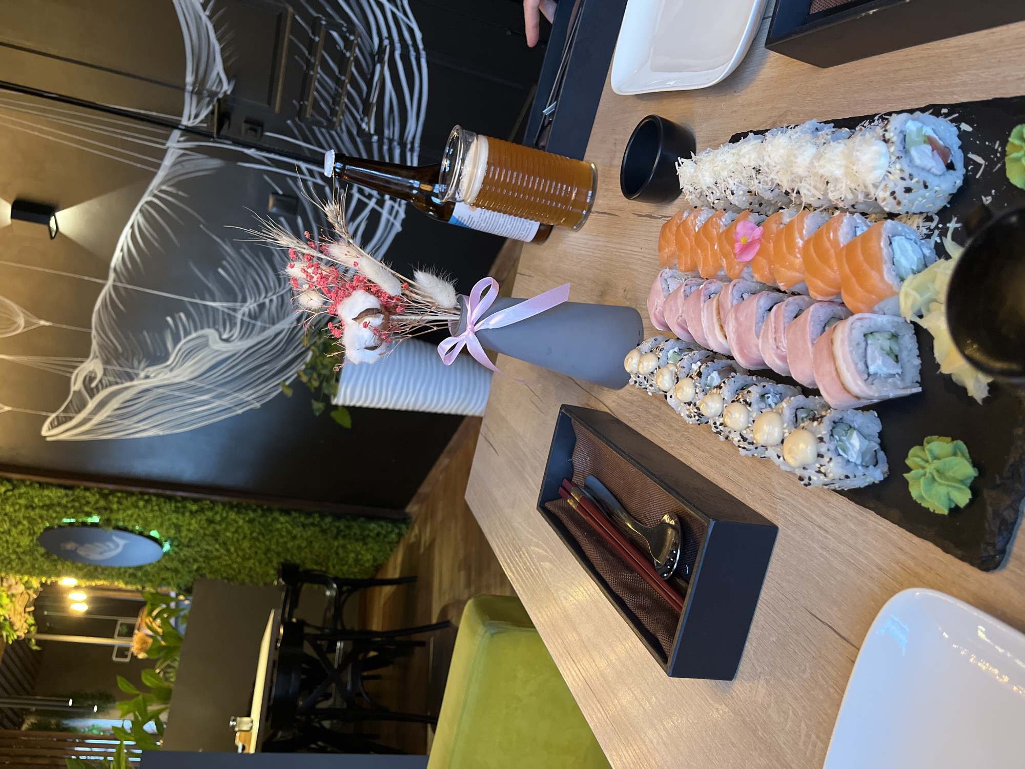 This media file captures a top-down view of a dining table laden with sushi and beverages in what appears to be a restaurant in Izhevsk, Russia.
The primary subjects are various types of sushi rolls, neatly arranged on a black slate-like platter. There are rolls topped with what looks like shredded coconut or cheese, rolls with slices of salmon, rolls wrapped in a light pink fish or meat, and rolls topped with creamy sauce and black sesame seeds. Alongside the sushi are servings of bright green wasabi and light yellow pickled ginger.
Accompanying the meal is a bottle of beer, from which a glass has been poured, showing a golden amber liquid with a foamy head. The beer bottle has visible text, including "БАРНХАУС" (Barnhaus), "БОГЕМСКИЙ ПИЛСНЕР" (Bohemian Pilsner), "КЛАССИЧЕСКИЙ ЛАГЕР" (Classic Lager), and "СВЕТЛОЕ" (Light).
The table is made of light wood. Tableware includes a black box containing red chopsticks and a small spoon, a small black bowl, and white plates. A decorative grey vase tied with a pink ribbon holds a bouquet of dried flowers, featuring white cotton pods, pink dried blooms, and wheat-like stalks, adding a natural touch to the setting.
The background reveals a modern restaurant interior. One wall is dark with white, abstract, and possibly floral or wave-like linear drawings. Another wall is covered with artificial green foliage, creating a vibrant texture. Accent lighting fixtures are visible on the dark wall. Chairs, other tables, and glimpses of a wider dining area can be seen in the periphery. The general ambiance suggests an evening setting, given the reliance on artificial lighting. A person's hand is subtly visible near the beer bottle, indicating the presence of diners, though no faces are shown.