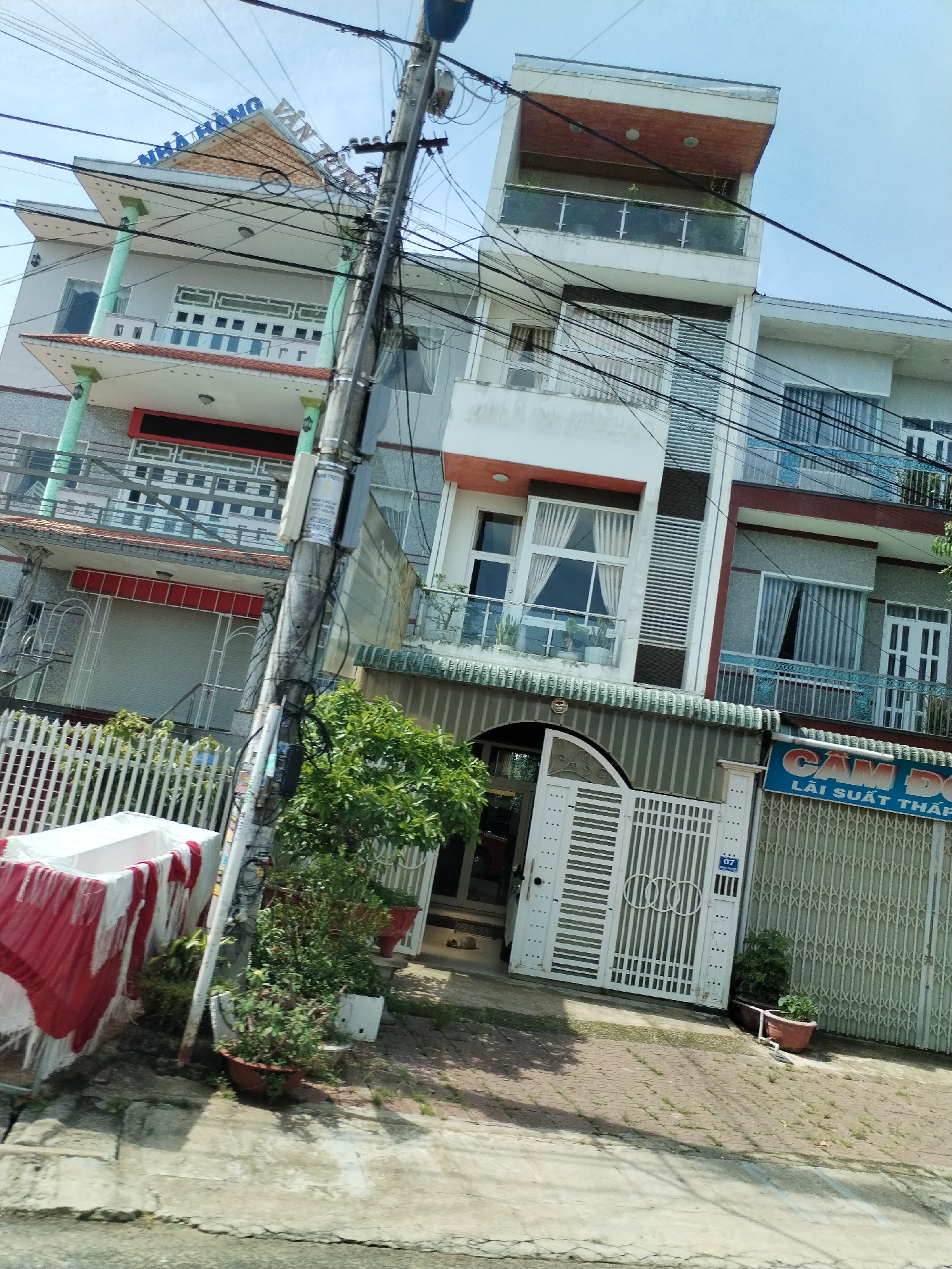 The image depicts a street scene in Bờ Y, Vietnam. Three multi-story buildings are visible, each with distinct architectural styles. The building on the left features a sign in Vietnamese that reads "NHÀ HÀNG VĂN TÚ." A building in the center has a white gate with a small blue sign reading "07" on it. Another building on the right has blue signs with text in Vietnamese reading "CAM DO LÃI SUẤT THẤP." Power lines run across the scene, obscuring the view. A utility pole stands in the foreground. Plant pots with greenery are placed near the buildings. A red and white object sits near a fence. The street surface appears uneven and patched.