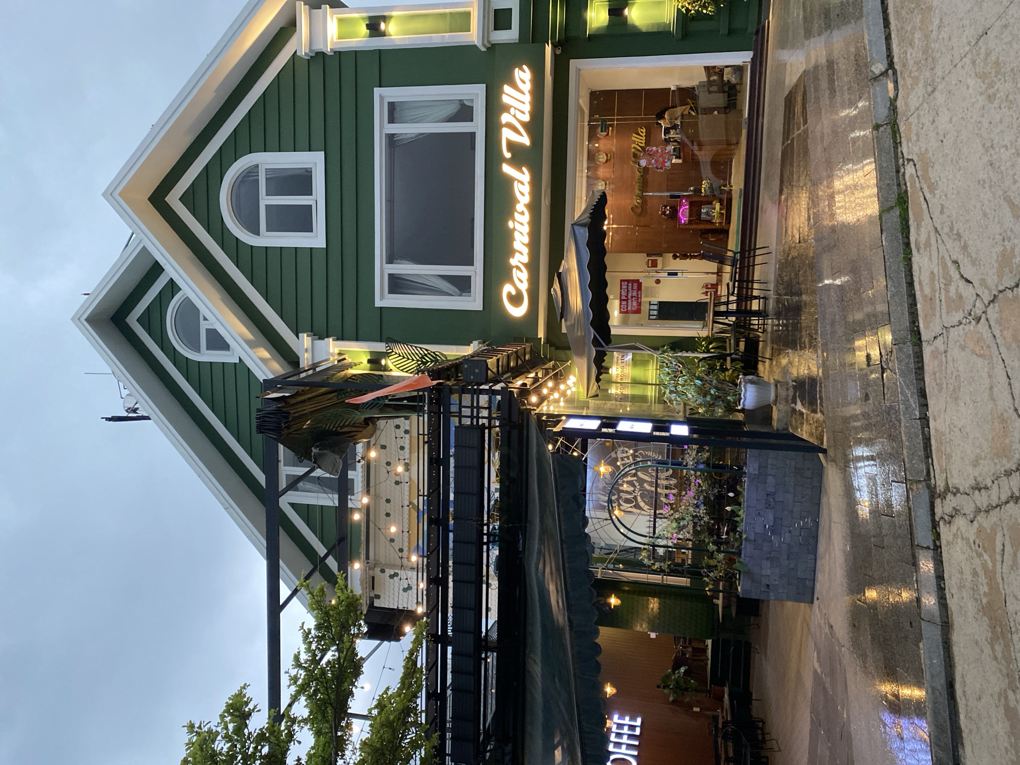 This image captures the exterior of a charming two-story building, likely a cafe or restaurant named "Carnival Villa," set in Da Lat, Vietnam, during dusk or early evening. The building features a distinctive design with green horizontal siding and crisp white trim, including decorative eaves and arched windows on its upper gables.
The main facade displays "Carnival Villa" in bright, illuminated script lettering, indicating it's open for business. Below this sign, there's a large rectangular window and a glass entrance, offering a glimpse into the interior. An outdoor seating area extends from the building, featuring dark tables and chairs. This area is adorned with festive string lights that cast a warm glow, enhancing the inviting atmosphere. A dark-colored awning provides partial cover, and potted plants add greenery to the space.
The ground in front of the building and the adjacent sidewalk appear wet, reflecting the artificial lights from the building and street, suggesting it has recently rained or is currently drizzling. The sidewalk on the right shows cracks and some unevenness. No people are visible in the scene, leaving the outdoor area empty but prepared for patrons. Other visible text includes a partially obscured red sign inside the main window, possibly reading "CƠM PHÚC" with a phone number, and a partially cut-off "COFFEE" sign near the bottom right corner of the image. The overall impression is one of a cozy and well-lit establishment awaiting customers in the cool, possibly damp, evening air of Da Lat.
