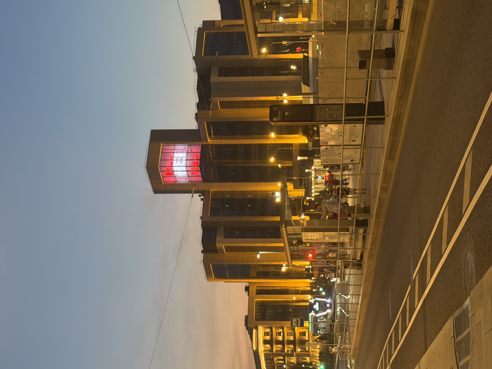 This detailed image captures an urban street scene in Moscow, Russia, at dusk, specifically along **Leningradsky Prospekt, in the vicinity of VTB Arena Park**.
The sky transitions from a deep blue overhead to a soft orange and pink gradient near the horizon, indicating twilight. Dominating the background are several multi-story modern buildings. A central, prominent building features a large, internally illuminated red square on its upper section, enclosing a bold white Cyrillic "В" (Ve) logo, strongly suggestive of VTB Bank. Adjacent to this, other modern office buildings exhibit dark glass facades accented by distinctive, brightly glowing vertical gold-yellow illuminated strips or frames. To the far left, an older building with classic architectural details is also brightly lit with warm yellow lights.
In the mid-ground, a wide asphalt road is visible. The foreground features a section of a street with white dashed lines on the left, indicating a designated lane. Parallel to the road, a modern, sleek white tram with illuminated turquoise accents and headlights on is seen moving towards the left. To the right, a tram stop is present, delineated by metal railings. This stop includes a tall, black information stand, partially obscured, displaying a tram icon and what appears to be a route number "1e" along with a small map. Several pedestrians are scattered across the tram platform and adjacent sidewalks, some waiting at the stop, others walking, appearing as indistinct figures in the warm evening light. The entire scene is well-lit by numerous streetlights emitting a warm yellow glow, complementing the illuminated buildings and visible traffic lights (both red and green). Overhead electrical lines are discernible against the fading sky. The overall atmosphere is that of a bustling urban environment transitioning from day to night.