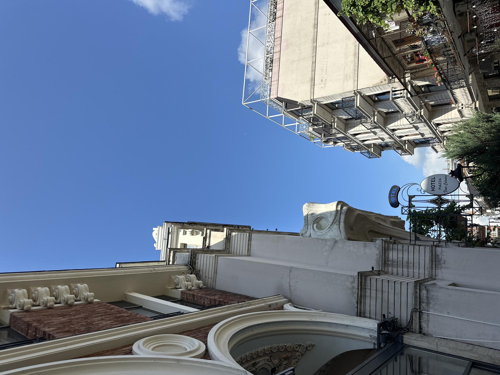 The image shows a low-angle view of buildings against a bright blue sky with some clouds. The architecture features decorative elements like intricate white moldings on the lower building and balconies with ornate railings on the upper buildings. The sunlight suggests it is daytime. One building has a sign that reads "HOTEL PIZZA Four Seasons," indicating a hotel or restaurant is present. The scene appears to be an urban environment, likely a street in Batumi, Georgia, given the architectural style and the mention of "Four Seasons." There are no people visible in the image, and no specific activity is taking place. The overall impression is one of a warm, sunny day in a city with a mix of classic and modern architecture.