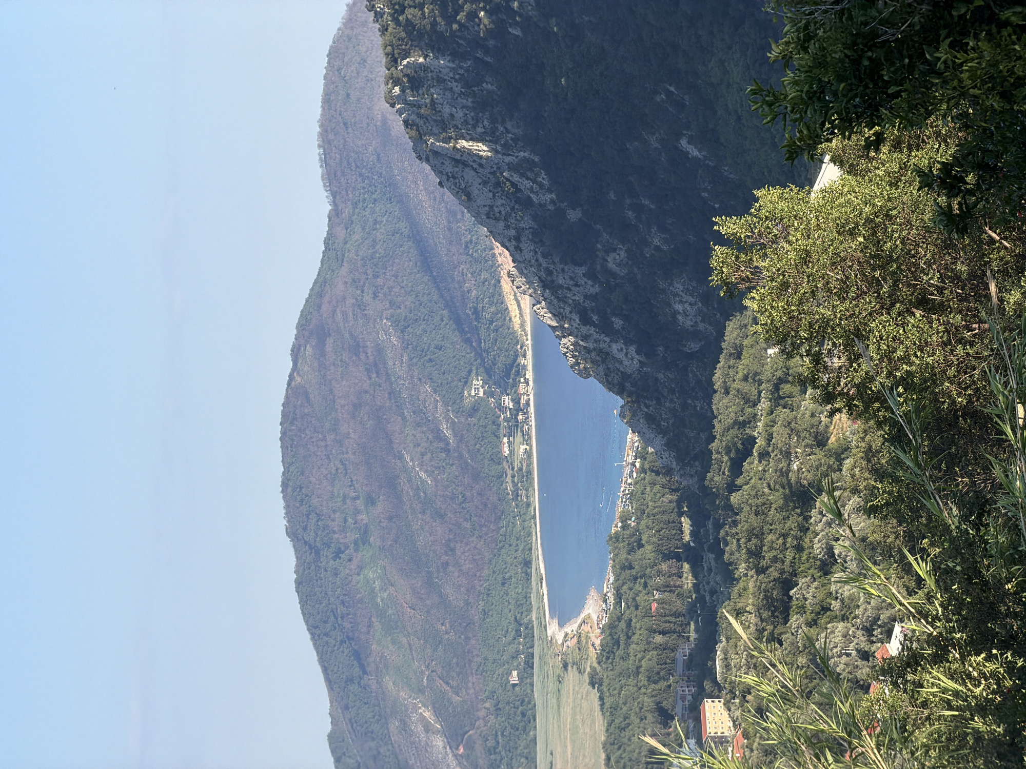 The image captures a scenic overlook of a bay and surrounding landscape in Androvići, Montenegro. The foreground is dominated by lush green vegetation, including tall reeds and dense trees, with glimpses of buildings nestled among them. As the view extends backward, a crescent-shaped bay with calm, blue water comes into focus, bordered by a sandy beach lined with a scattering of people and what appears to be a road or promenade. Beyond the bay, gentle hills covered in greenery and some areas of dry, discolored vegetation rise up, eventually giving way to steeper, rocky mountains in the background. The sky is clear and a pale blue. The scene suggests a tranquil coastal setting with a mix of natural beauty and human settlement.