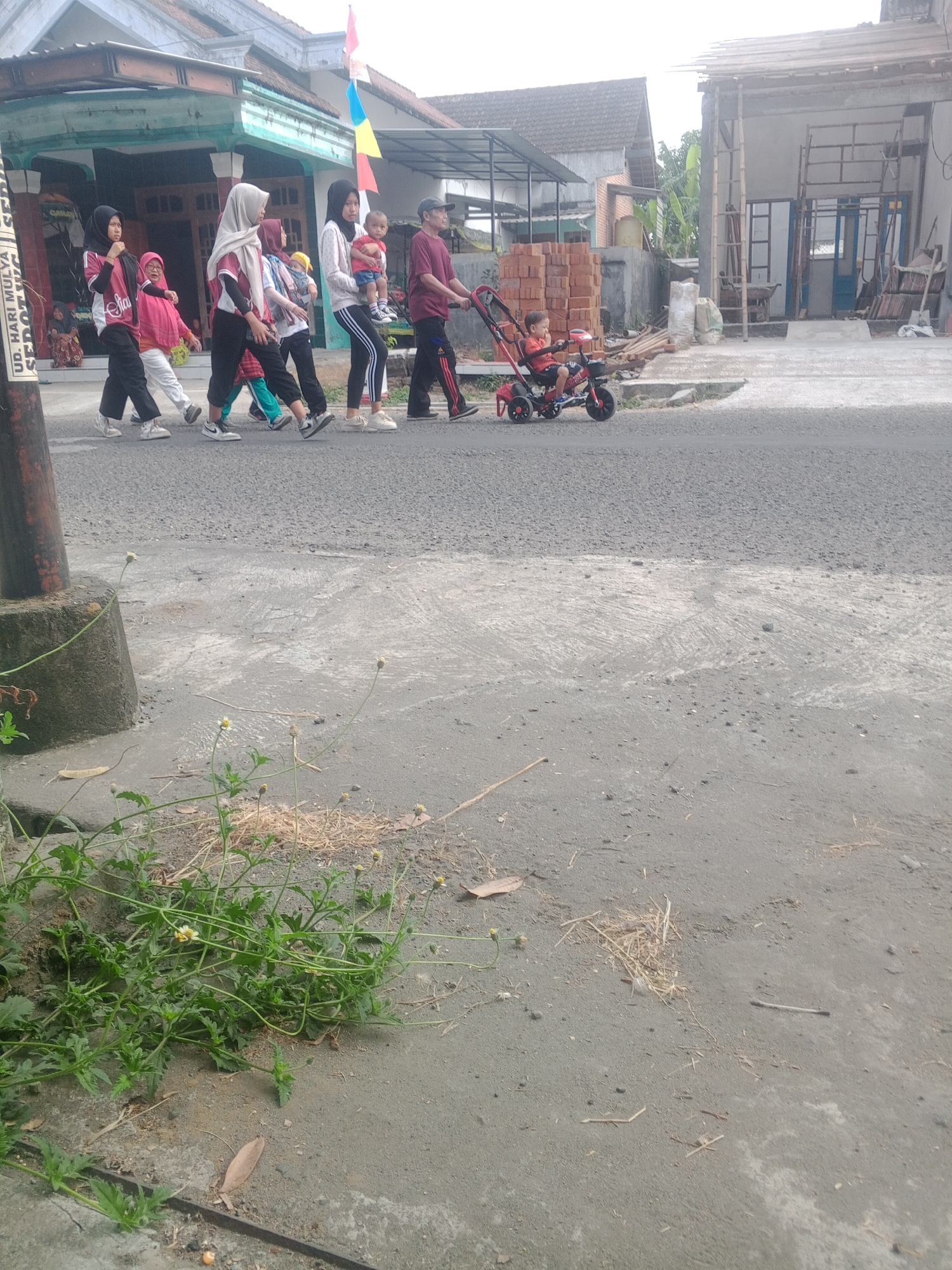 In the city of Lembah, Indonesia, a group of people walks along a paved road. A woman in a maroon shirt and black pants pushes a red stroller containing a child wearing a red shirt. Behind her, a woman in a white shirt and black pants carries a baby. To their left, a woman in a black hijab and pink shirt walks with a young child in a pink shirt and white pants. Another woman in a white hijab and black pants walks beside them, and another woman in a black hijab and white shirt is further ahead. Several buildings are visible in the background, including a house with a green trim and a structure under construction. The road is lined with a concrete sidewalk and patches of grass and weeds.