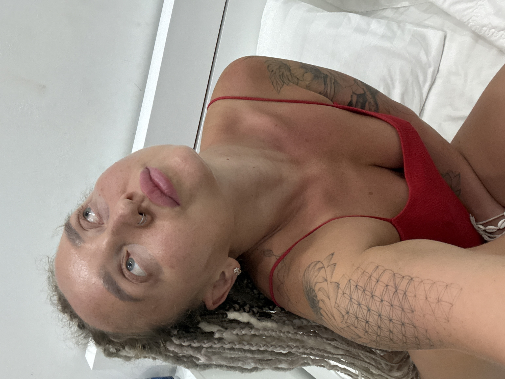 A woman with grey braids, a nose ring, and tattoos on her left arm and right shoulder is looking upwards. She is wearing a red swimsuit and sitting on a white bed with a white pillow. The room appears to be a bedroom with a white wall and headboard.