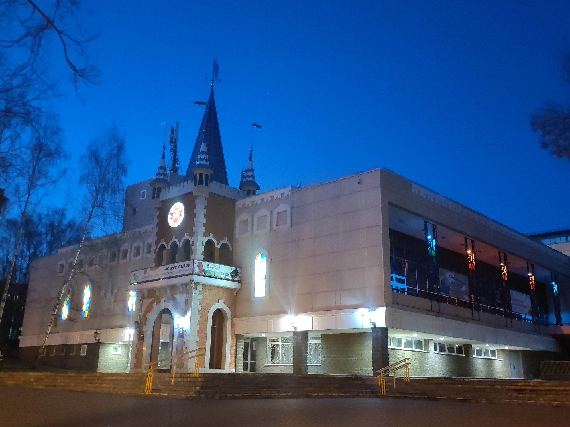 This night-time or deep twilight image captures a distinctively styled building in Izhevsk, Russia, under a clear, dark blue sky. The architecture blends castle-like features with more modern elements.
The central part of the building, illuminated by artificial lights, boasts a facade resembling a fortress or fairy tale castle, complete with spires, crenellated battlements, and arched window and door frames. A large, circular, glowing emblem with what appears to be a stylized star or sun is prominent on the main tower. Several spires top the roofline, one featuring a weather vane shaped like a bird. Ornate stained-glass windows, glowing with vibrant colors, are visible on the lower left section of this older-style facade. Steps with yellow handrails lead up to the main entrance.
To the right, the building shifts to a more contemporary design with expansive, multi-paneled glass windows on the upper floor, revealing internal displays or banners. The lower part of this section features a base of dark, textured stone and smaller, horizontally aligned windows.
Bare trees, likely indicating autumn or winter, frame the left side of the structure, silhouetted against the dark sky. No people are visible in the scene.
Visible text on a banner above the main entrance reads "НОВЫЙ СЕЗОН" (New Season). Below it, another banner displays "ВЕСЬ СЕЗОН" (All Season) and partially legible Russian text including "КУЛЬТУРА" (Culture) and a website address, suggesting the building serves as a cultural or entertainment venue.