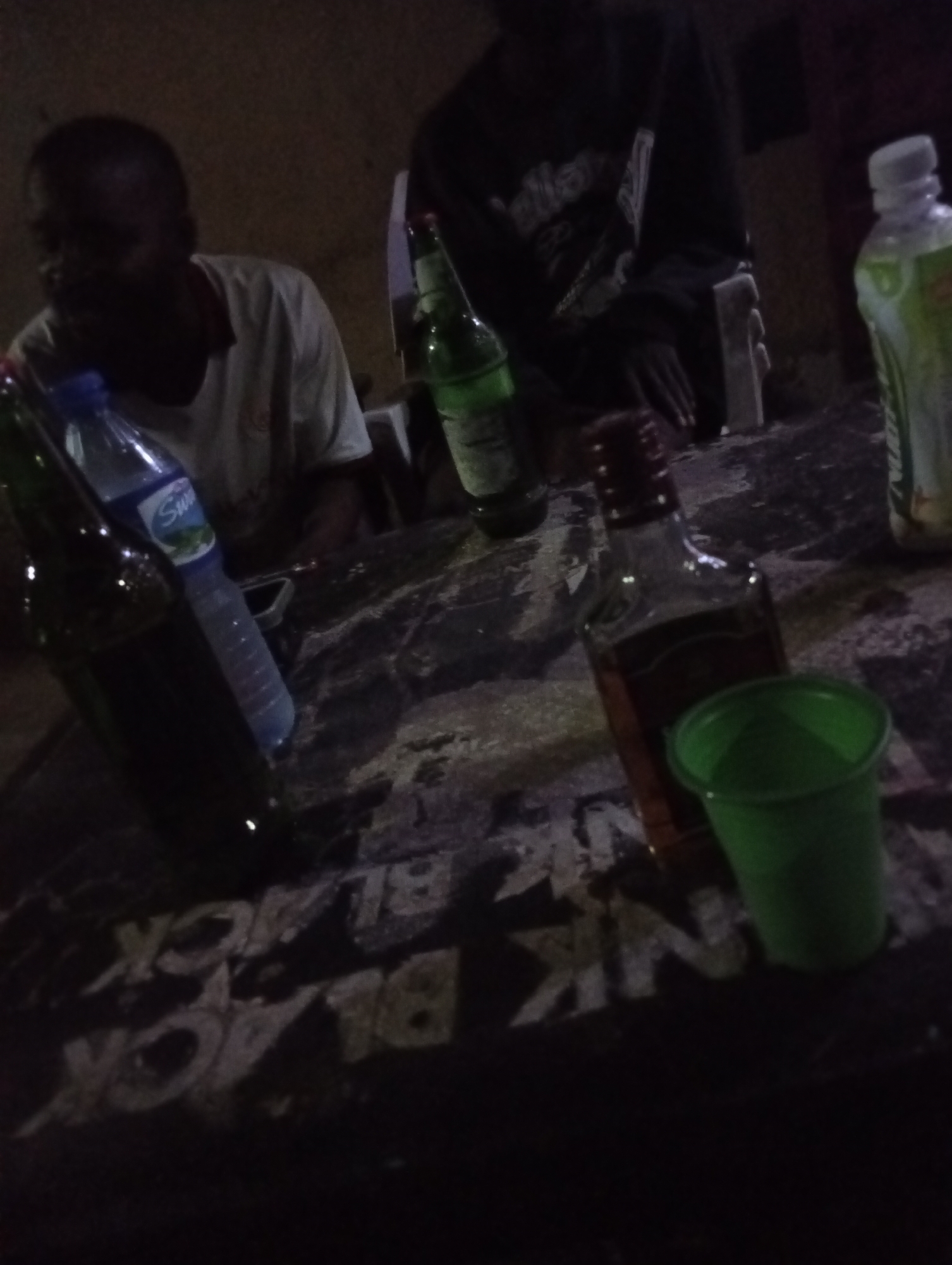 The image depicts a dimly lit scene likely in a social gathering setting in Jos, Nigeria, suggested by the presence of alcoholic beverages and the casual attire of the individuals present. Two people are partially visible: one wearing a white t-shirt with a red neckline and another wearing a black t-shirt with white writing. Several bottles are arranged on a table with “BLACK JACK” printed on it. The bottles include a bottle of Star beer, a bottle of water, a bottle of spirit, and a bottle of a green-colored liquid, along with a green plastic cup.