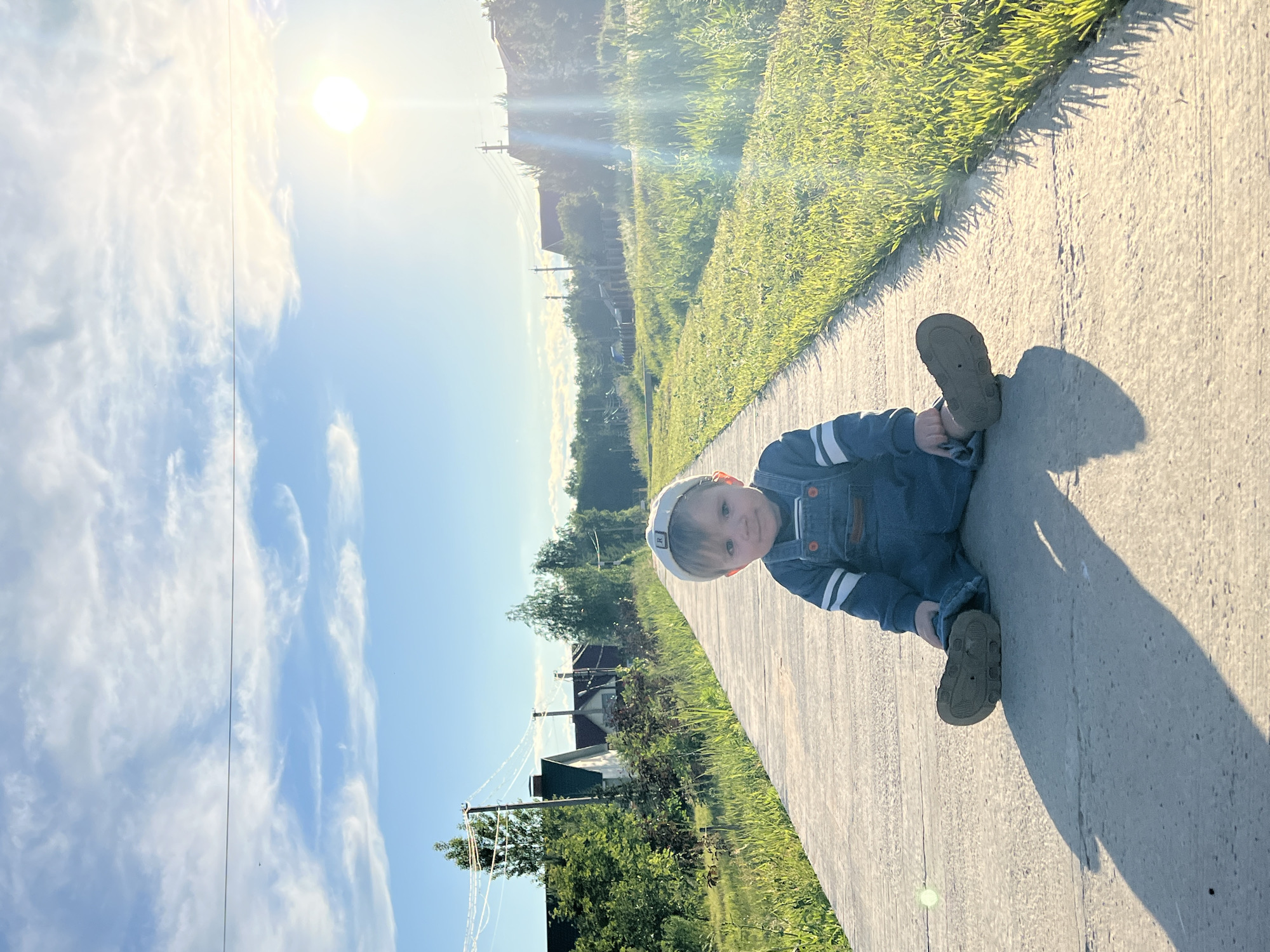 A young child, appearing to be a toddler, is seated on a concrete sidewalk on a bright, sunny day in Noviy, Russia. The child wears a light-colored baseball cap with a subtle white letter or logo (possibly an 'R'), a blue long-sleeved shirt with white stripes on the sleeves, and blue denim overalls, along with dark-colored shoes. The child looks directly at the camera with a calm, curious expression.
The scene is an outdoor, rural or suburban landscape. A concrete path stretches diagonally across the foreground and midground. To the right of the path is a strip of vivid green grass, brightly lit by the sun. In the background, beyond the grass, are various trees, power lines, utility poles, and several distant houses or buildings.
The upper left portion of the image features a clear blue sky adorned with soft, wispy white clouds. A brilliant sun, situated in the upper left, casts a strong, warm light across the scene, creating lens flare and long, distinct shadows, including the child's own shadow stretching across the sidewalk. The low angle of the sun suggests it is either early morning or late afternoon.