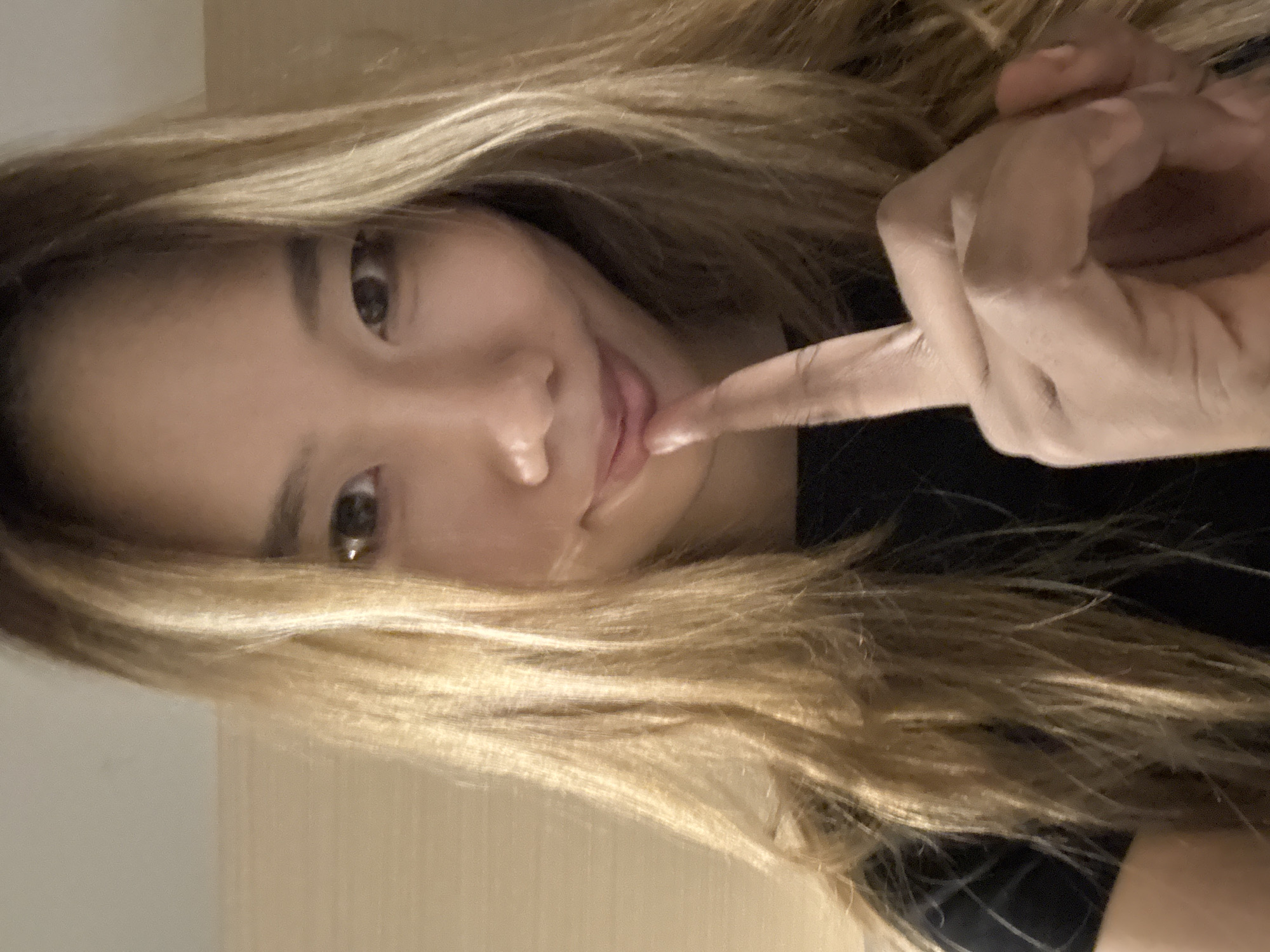 A close-up, waist-up shot shows a young Asian woman with blonde hair, wearing a black t-shirt. She has her right index finger to her lips, in a "shushing" gesture. Her eyes are dark, and she has a subtle smile. The background appears to be a wall, possibly in a room. There is no indication of the location being in Yuzhong District, China.