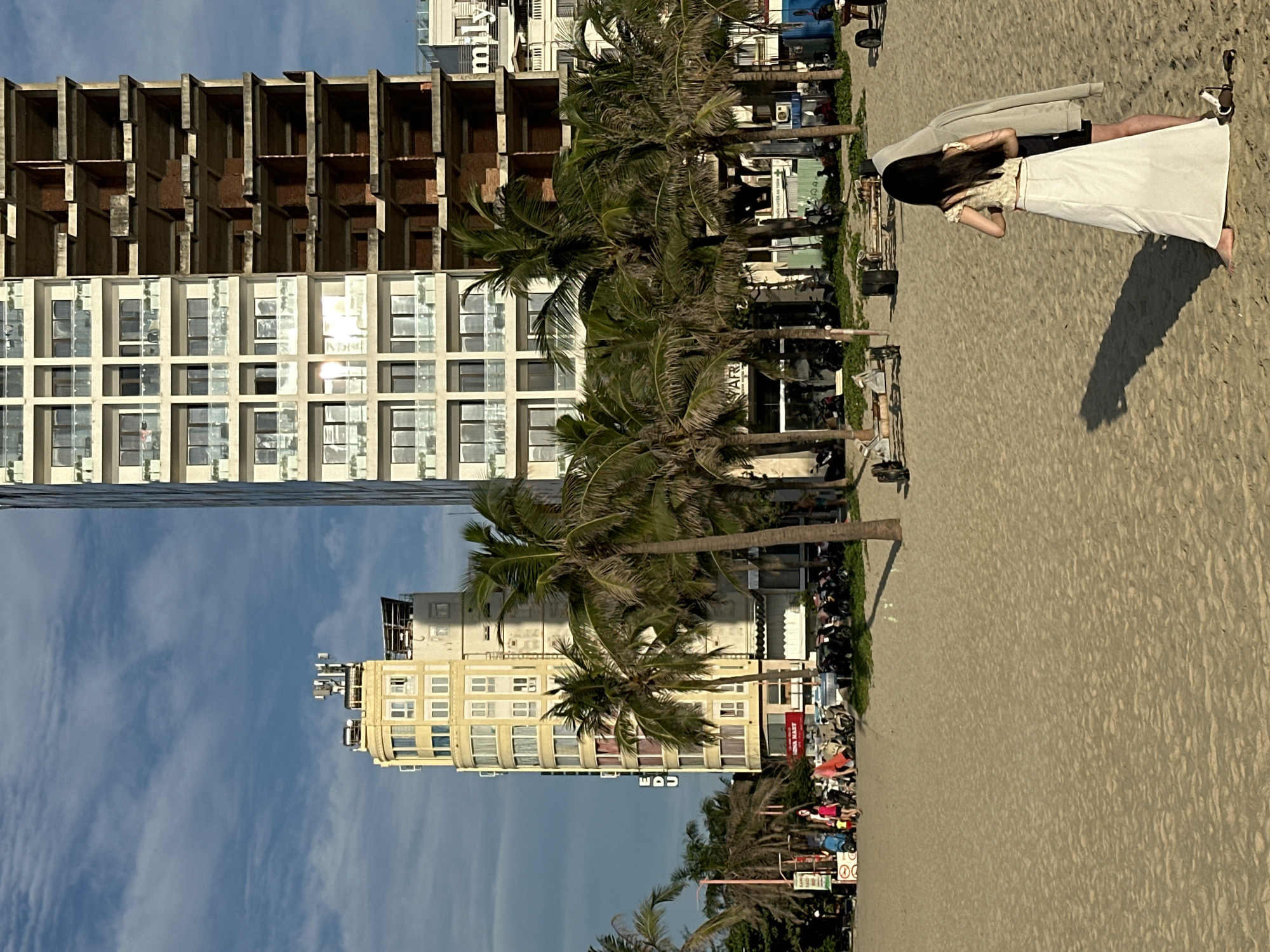 The image depicts a scene on a sandy beach in Da Nang, Vietnam, with a backdrop of urban development. In the foreground, two individuals are standing on the beach, facing away from the camera, with one person wearing a long white skirt and the other obscured by a hooded garment. The beach is textured with sand ripples.
Behind them, a row of palm trees lines the edge of the beach, separating it from a street and several buildings. Several motorcycles are parked along the street. Two prominent buildings dominate the midground and background. The building on the left is a yellow, multi-story structure with numerous windows and a rounded facade, featuring antennas on its roof. To its right is a much taller, unfinished modern building under construction, showing exposed concrete floors and brickwork on its right half. Further to the right, another building with balconies is visible.
The sky above is a clear blue with scattered white clouds, and the sunlight casts shadows on the sand, indicating it is likely late afternoon or early morning. The overall impression is a contrast between a natural beach environment and the ongoing urban expansion.