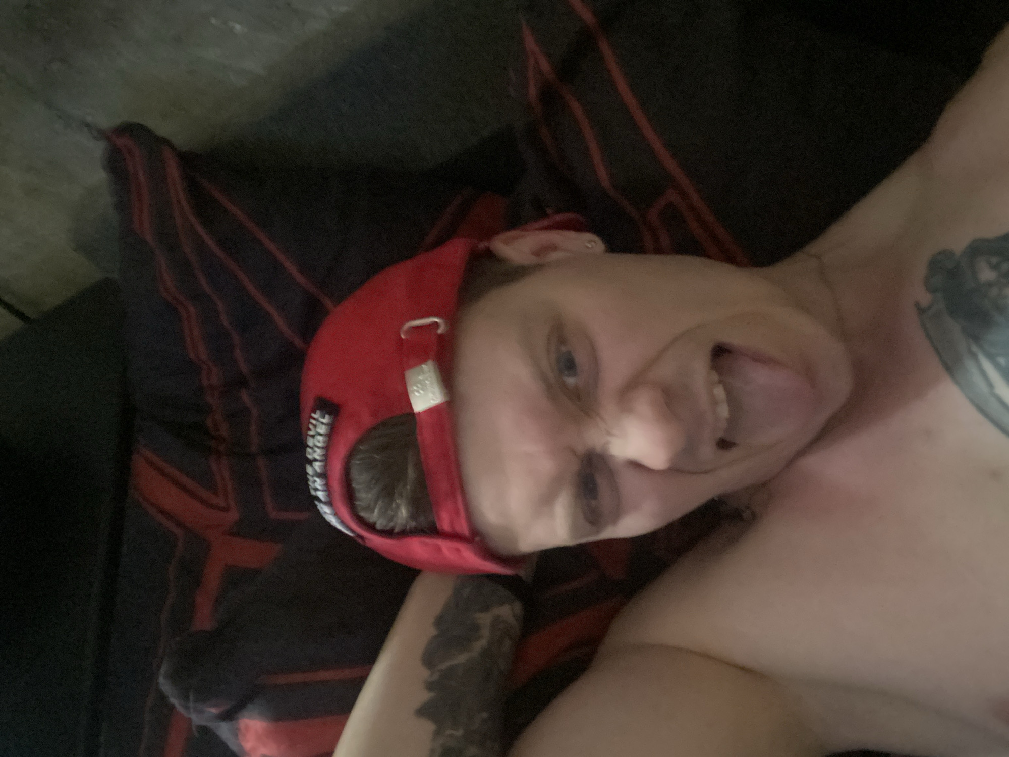 In the foreground of the image, a person with a red baseball cap is shown from the chest up, lying on a surface with a black and red striped pattern. The person is sticking their tongue out and making a playful or mischievous facial expression. A tattoo is visible on their left arm, and another tattoo is partially visible on their chest. The background consists of the striped surface, which appears to be bedding or upholstery. The lighting is dim, suggesting an indoor setting, possibly a bedroom. The provided crops offer closer views of the person's face, tattoos, and the textured background. No identifiable landmarks or elements specific to the city of Samara, Russia, are visible in the image.