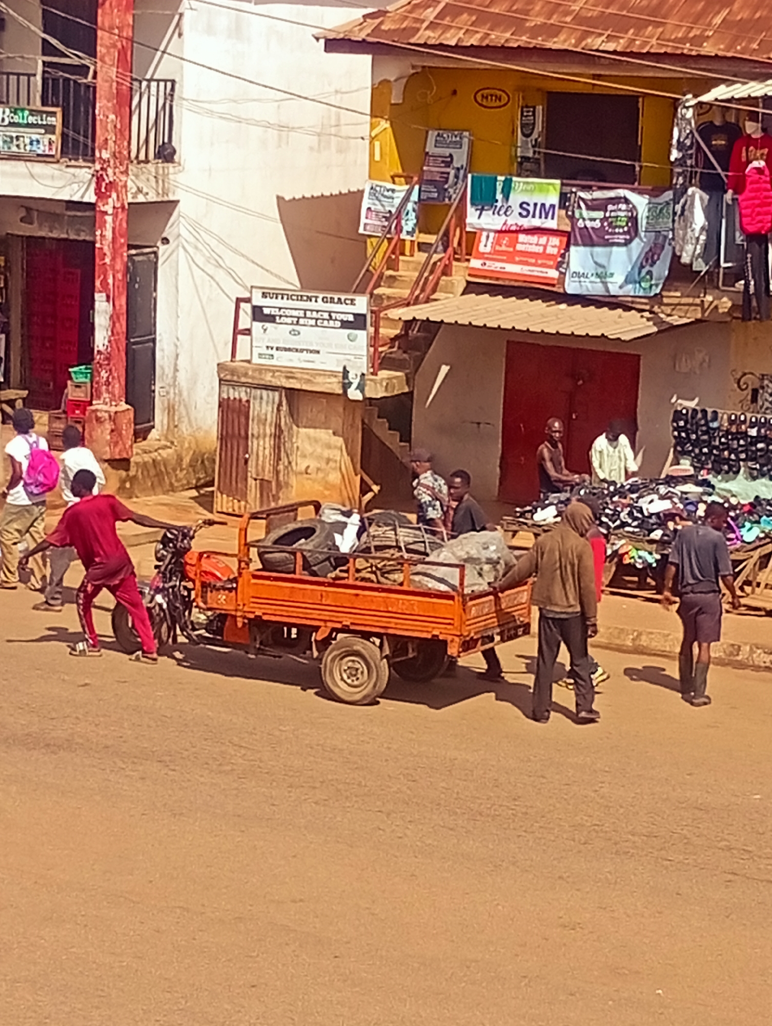 In Bukuru, Nigeria, a street scene unfolds with a focus on a bright orange motorized tricycle cart laden with tires and other items. A person in a red outfit appears to be interacting with the tricycle. Behind them, two individuals are walking away from the camera. Further in the background, several people are engaged in activities around market stalls. One stall displays a variety of footwear, with a person behind the counter. Another stall features clothing and advertisements. Buildings with corrugated iron roofs and balconies line the street, with a prominent yellow structure housing what appears to be a business. The street is paved, and the overall atmosphere suggests a bustling commercial area.