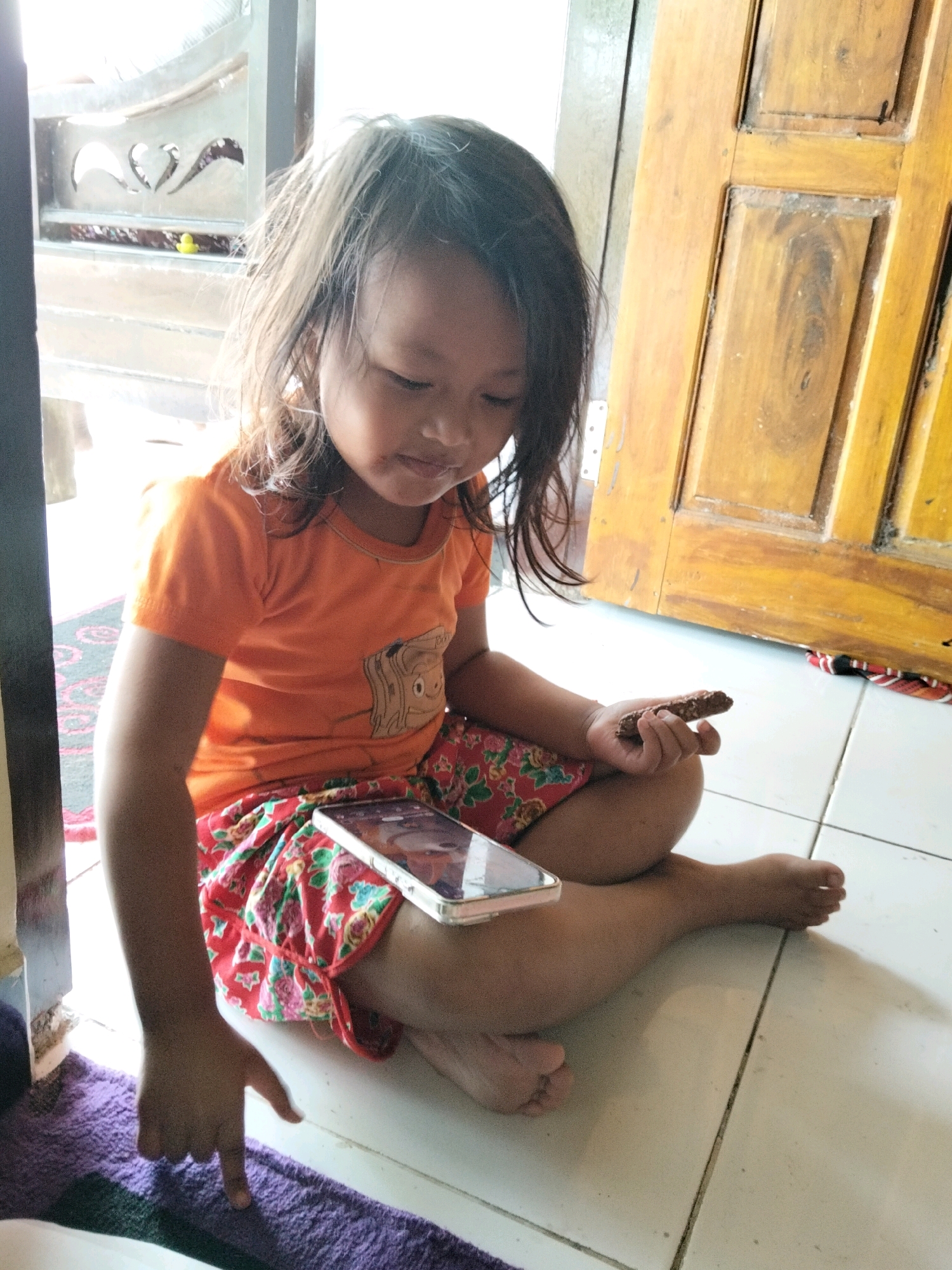 A young girl with long, dark hair sits cross-legged on a tiled floor in a room with natural light. She is wearing an orange t-shirt with a cartoon character graphic and a red skirt with a floral print. Her left hand rests on her lap, while her right hand holds a small, brown, oblong object that appears to be a snack or a toy. A smartphone lies on her right leg, its screen illuminated. In the background, a wooden door with carved panels is visible, along with what looks like an ornate metal gate. The lighting suggests it could be daytime. The girl's gaze is directed downwards, and her expression is neutral. The scene appears to be indoors, possibly in a home, given the domestic setting and the presence of a child.
