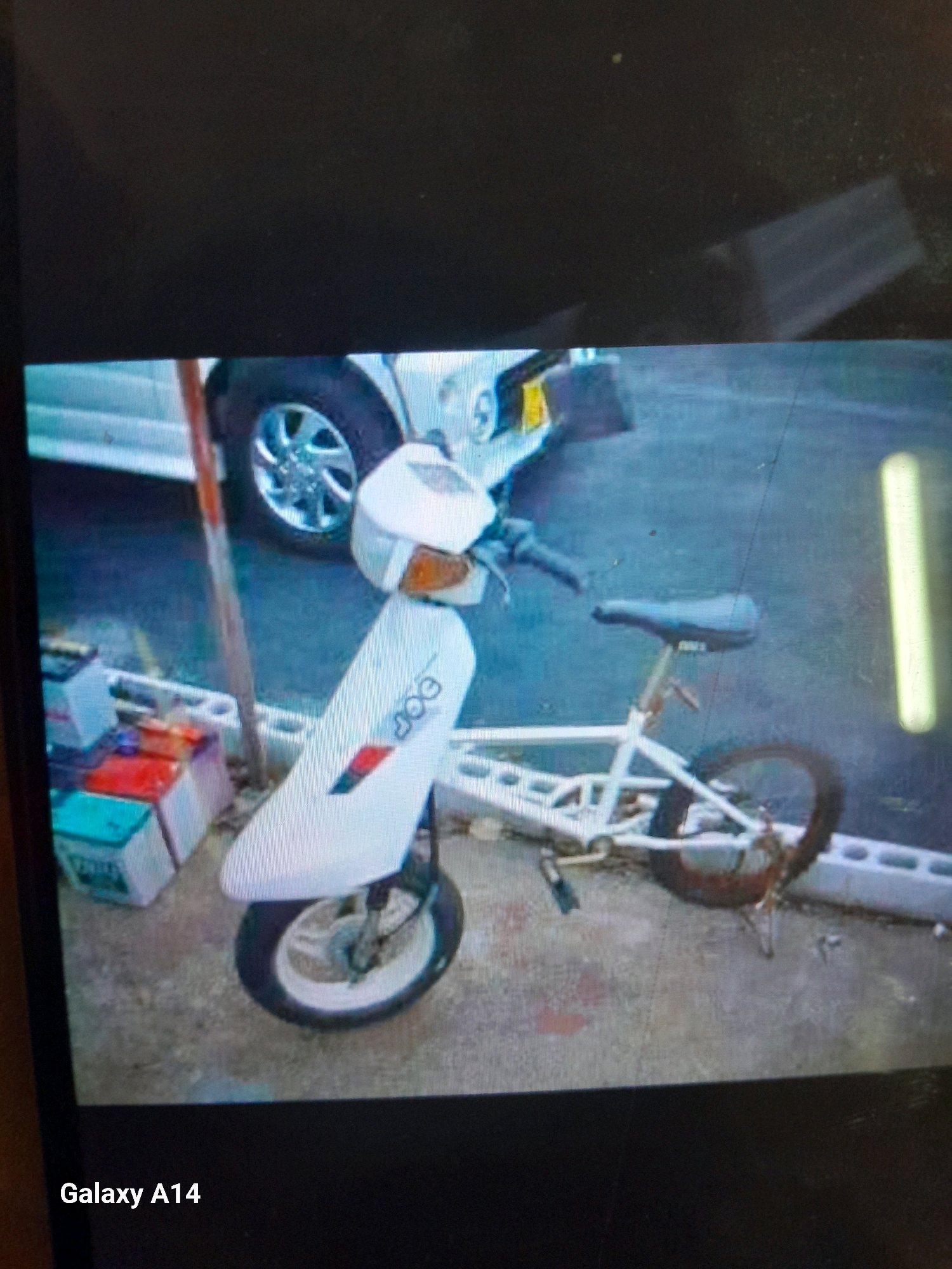 The image shows a white, custom-built vehicle that resembles a cross between a scooter and a bicycle, parked on a paved surface in what appears to be an outdoor setting. The vehicle has a prominent white body with a sleek, aerodynamic design, and is equipped with a seat, handlebars, and wheels. The front wheel is smaller than the rear wheel, and the vehicle has a kickstand. The background includes a portion of a vehicle, possibly a white SUV, with a visible tire and rim, and a vertical pole or post. There are also several stacked items in the background that appear to be batteries or storage containers, some of which are colored red, blue, and green. The ground is asphalt or concrete, with some debris. The text "Galaxy A14" is visible in the lower left corner, indicating the device used to capture the image. There are no people or specific geographical markers to definitively place the location in Noord, Suriname.
