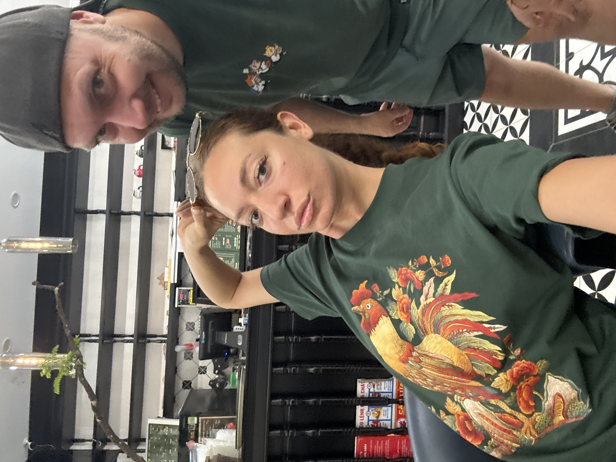 The image, a portrait-oriented selfie, features a woman in the foreground and a man behind her, taken indoors. The woman, with long brown hair and sunglasses perched on her head, wears a dark green t-shirt showcasing a vibrant graphic of a rooster surrounded by red and orange flowers. She holds the camera up, capturing the shot with a neutral to slightly serious expression. Behind her, partially visible, is a man wearing a dark cap and a dark green t-shirt with a small, indistinct graphic of cartoon characters. He looks towards the camera with a slight smile.
They are situated in what appears to be a cafe, restaurant, or shop. The background reveals modern interior design elements, including black and white geometric patterned tiles on a wall or floor, and dark shelving units. Contemporary cylindrical light fixtures and a decorative branch with foliage add to the ambiance. On the shelves, various products are displayed, some with visible Vietnamese text, including "LÊNH", "CA LU", "CA PHE" (coffee), and "TRÀ" (tea), indicating the type of establishment. The scene conveys a casual and relaxed atmosphere, likely during daytime.