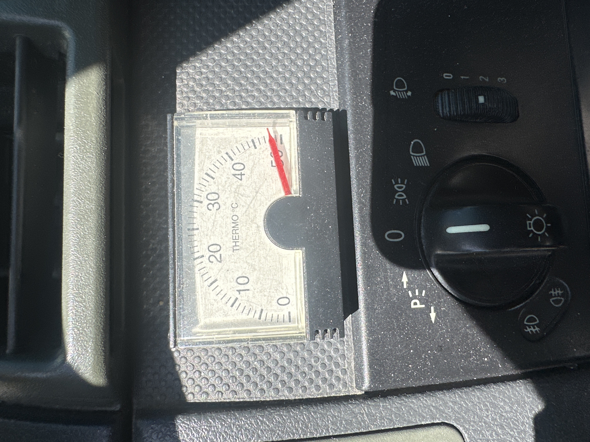 The image shows a close-up view of a vehicle's dashboard, focusing on a thermometer and control knobs. The thermometer is circular with a white face, marked with temperatures from 0 to 50 degrees Celsius. A red needle indicates the current temperature, pointing slightly above 40 degrees Celsius. Below the thermometer, there are several control knobs and symbols. One knob, to the left, has symbols that appear to represent parking brake engagement. To the right of this, there is a rotary switch with markings 0, 1, 2, and 3, likely for adjusting some vehicle function. Symbols indicating headlight functions are also visible. The dashboard material appears to be dark-colored plastic with a textured surface. The lighting suggests it is daytime, with a strong light source casting shadows on the dashboard. The location is unknown, city unknown.