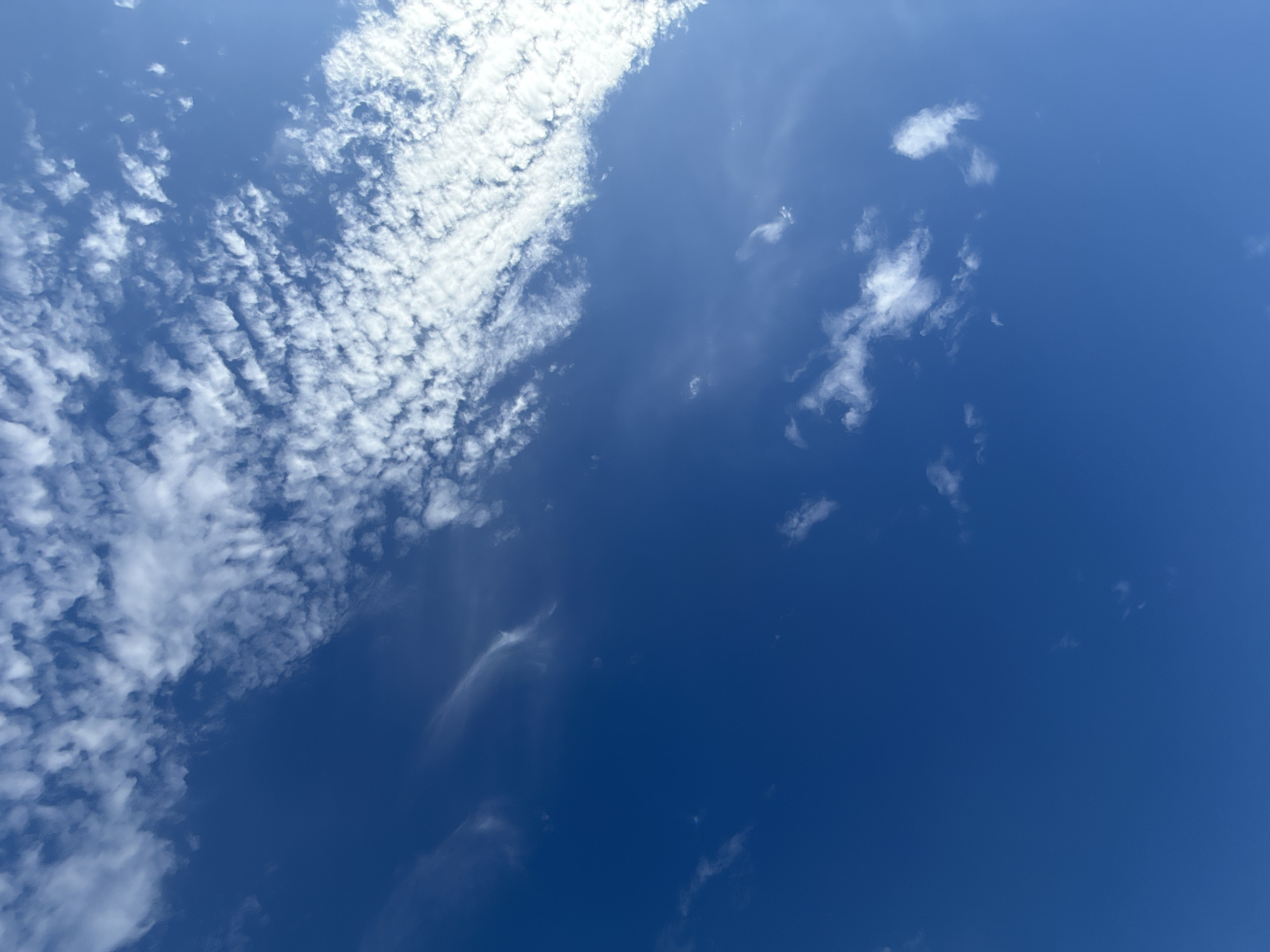 This is a daytime photograph of a bright blue sky with scattered white clouds. The clouds are wispy and varied in density, with a larger band of textured clouds stretching across the upper right portion of the frame. The lower portion of the image is predominantly clear blue sky with a few smaller, isolated clouds. The image appears to be taken from a ground-level perspective, looking upwards. There are no people, structures, or any other discernible objects or actions visible in the scene. This photograph is not specifically indicative of Bielany Wrocławskie, Poland, as it depicts a universal natural phenomenon.