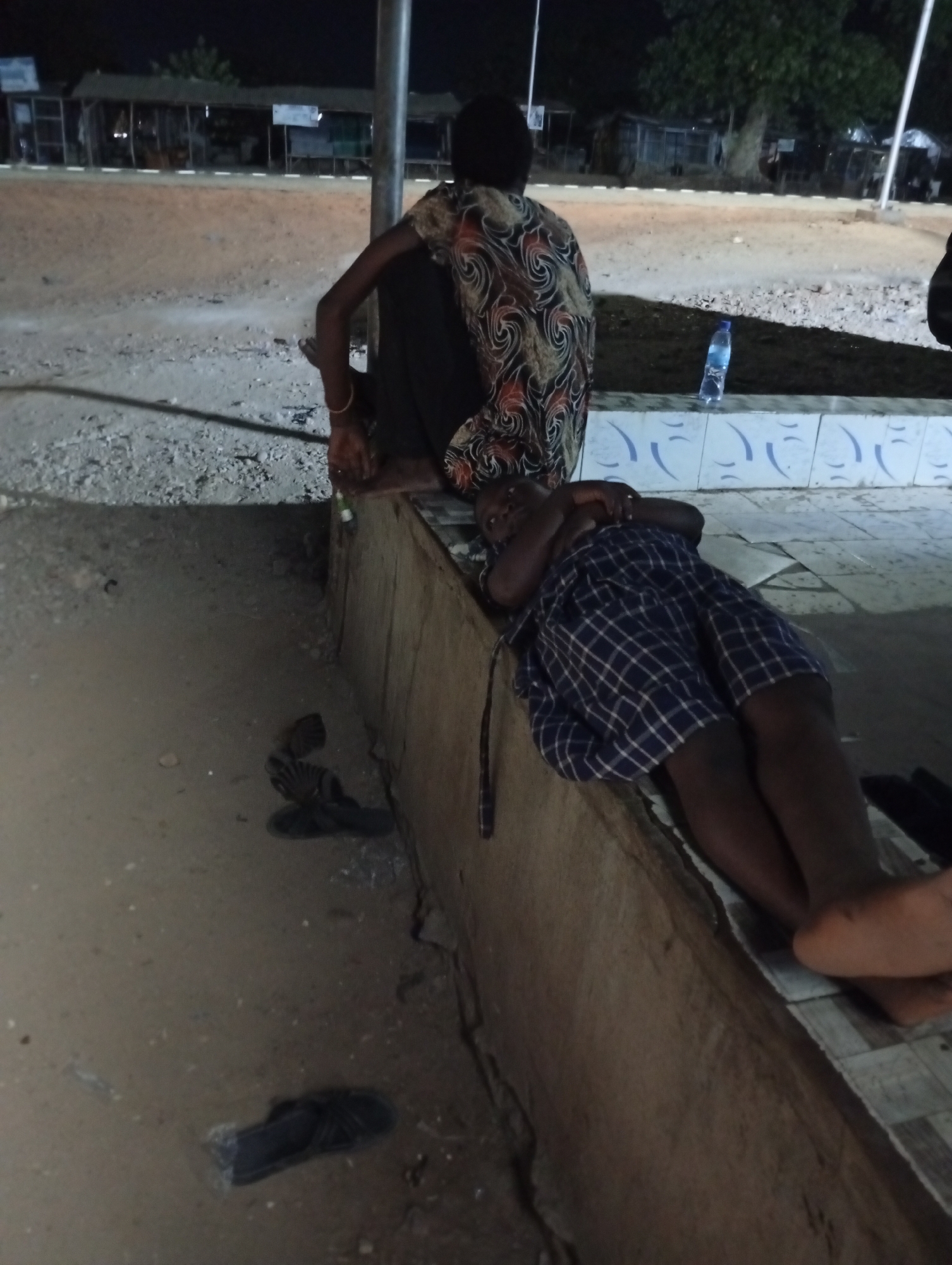 Two people are sitting and lying on a ledge. One person is sitting with their back to the camera, wearing a dark shirt with a multicolored pattern. The other person is lying down, wearing a blue and white checkered short and no shirt. There is a bottle of water on the ledge near the seated person. Several pairs of sandals or flip-flops are on the ground near the ledge. The location appears to be an outdoor public space at night, possibly in Adankolo, Nigeria. In the background are stalls and street lights.