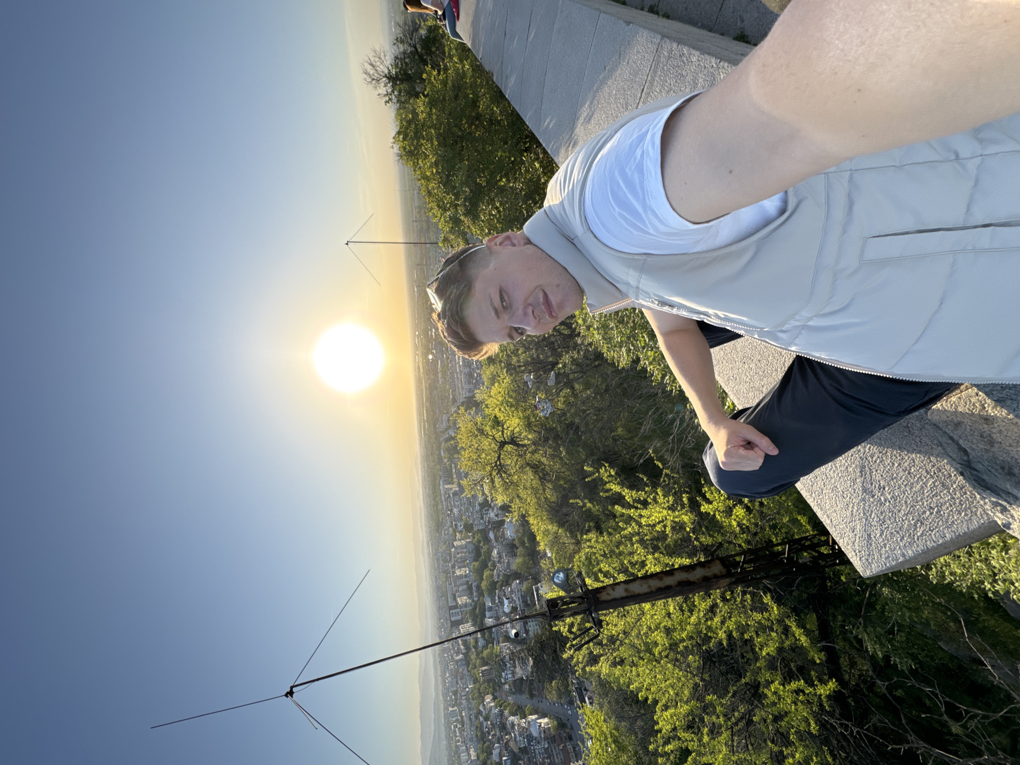 A man is taking a selfie at sunset overlooking the city of Plovdiv, Bulgaria. He is sitting on a stone structure, possibly a bench or wall, with green foliage visible below. The city skyline is in the distance, illuminated by the setting sun. Two antenna structures are visible in the foreground. The man is wearing a white t-shirt and a light-colored vest. He has short, blond hair. Part of another person is visible in the upper left corner of the image.