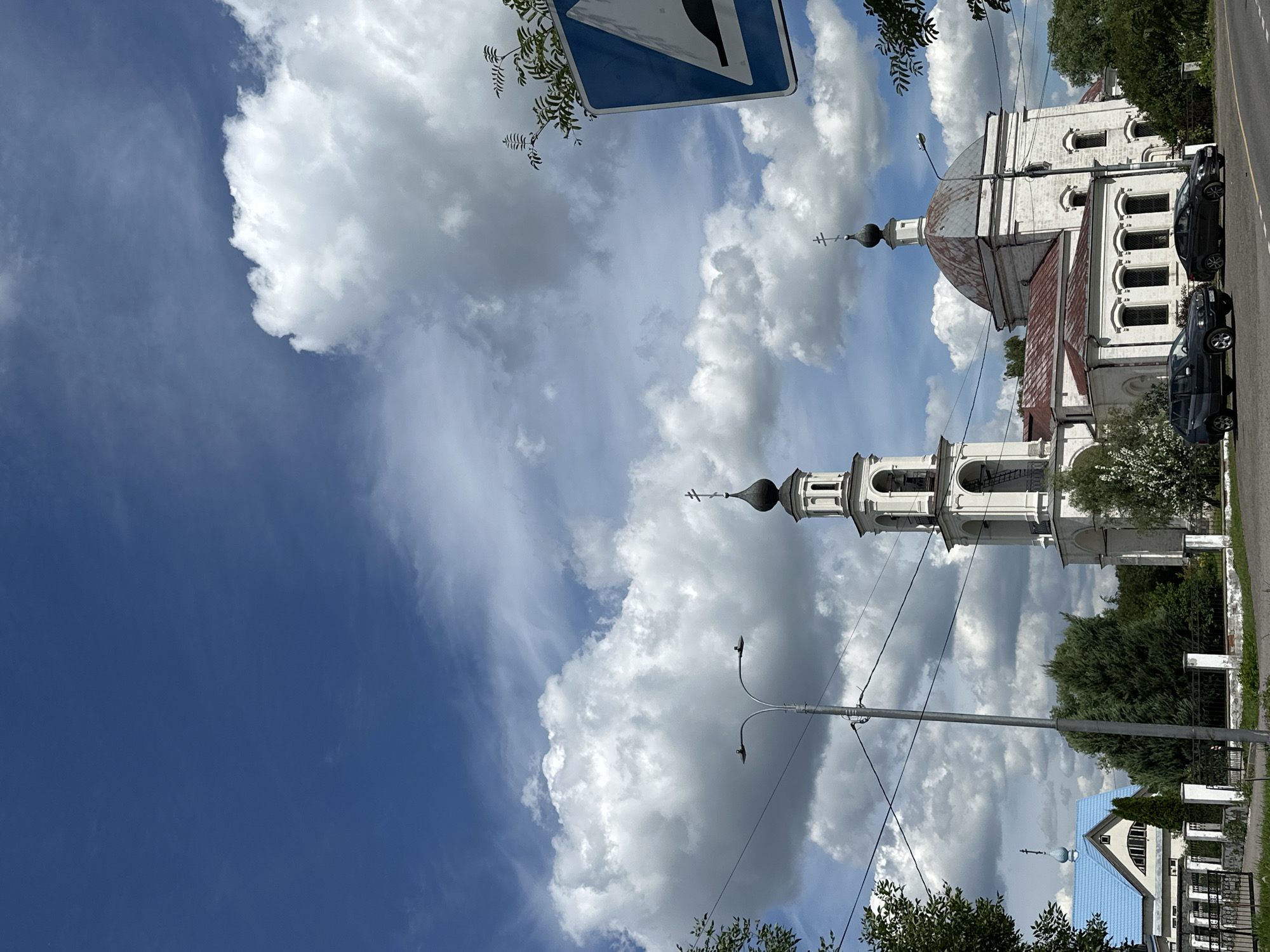 The image depicts a street view in Yerino, Russia, dominated by a bright blue sky with large, fluffy white clouds. In the center of the frame, a white church with a bell tower stands prominently. The church has a domed roof with a reddish-brown patina and a bell tower topped with a black spire and a cross. Adjacent to this church, to the left, is another building with a bright blue roof and a cross.
In the foreground, two dark-colored cars are parked on the side of a paved road. A street lamp with two lights is visible between the church and a line of trees. A blue and white road sign indicating a speed bump is positioned on the right side of the image, with branches of foliage encroaching on it. The scene is framed by green trees on the left and right edges, with a low fence in front of the buildings. The overall atmosphere is that of a sunny day in a semi-rural or suburban area.