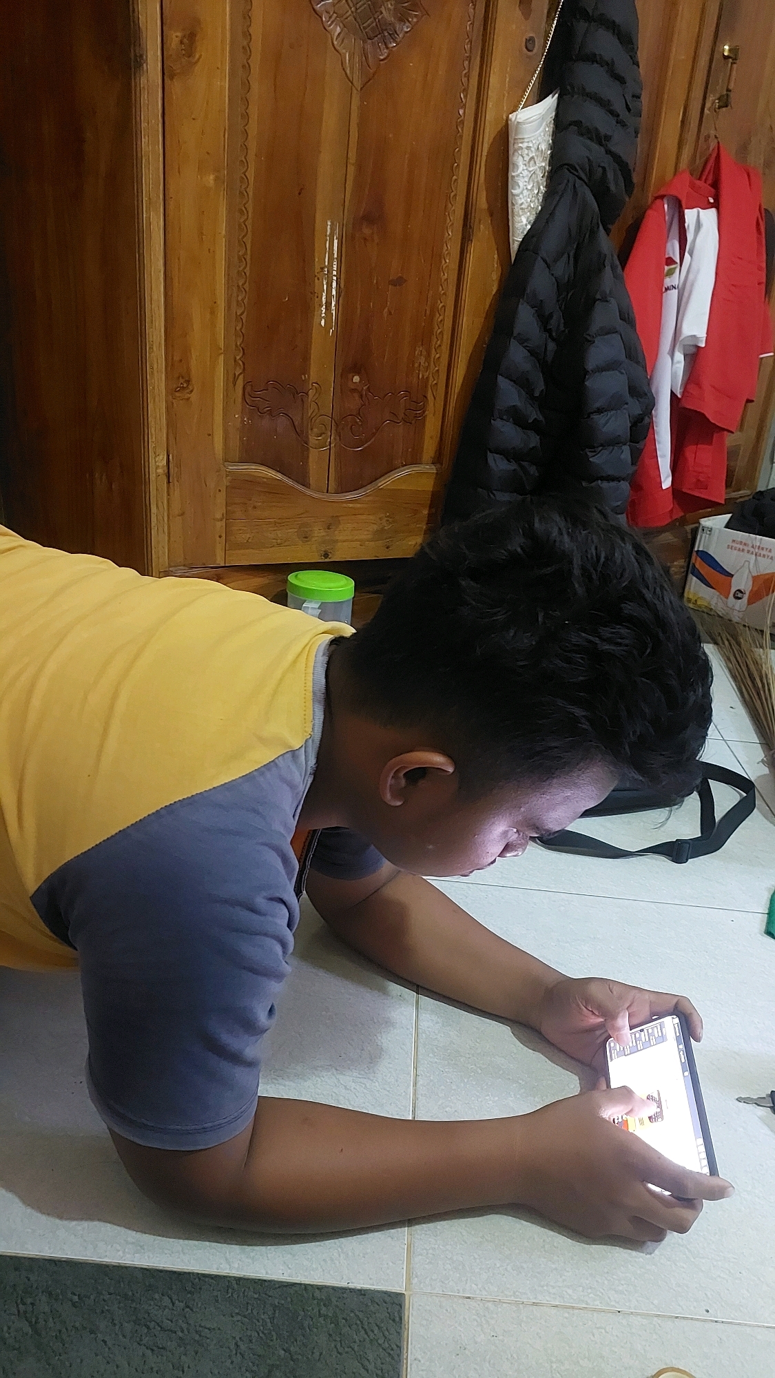 A person, likely a child or young adult, is lying prone on a tiled floor, engrossed in playing a game on a smartphone. The person is wearing a t-shirt with a yellow upper half and a blue-gray lower half. Their face is illuminated by the phone's screen. In the background, a large wooden wardrobe with intricate carvings stands against a wall. Two jackets are hanging on the wardrobe, one black and puffy, the other a red and white jacket. To the right of the wardrobe, a broom or similar cleaning tool is visible leaning against a wall. A green-lidded plastic container is placed on the floor near the wardrobe. The scene appears to be indoors, possibly in a residential setting in Sukowono, Indonesia, given the context provided.