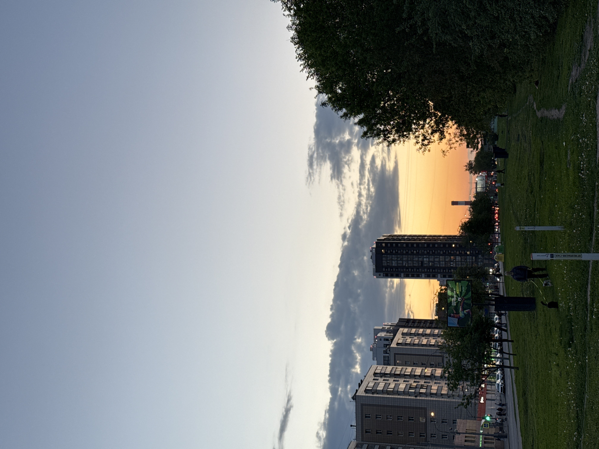 The image depicts a view in Moscow, Russia, during a sunset. The sky displays a gradient from blue to orange, with clouds situated around a tall residential building. In the foreground, a grassy area is visible, containing trees, a digital advertising billboard, utility poles, and a few individuals; one person, seemingly a male, is walking. Several buildings of medium and high rise stand clustered near the green area, marking an urban landscape.