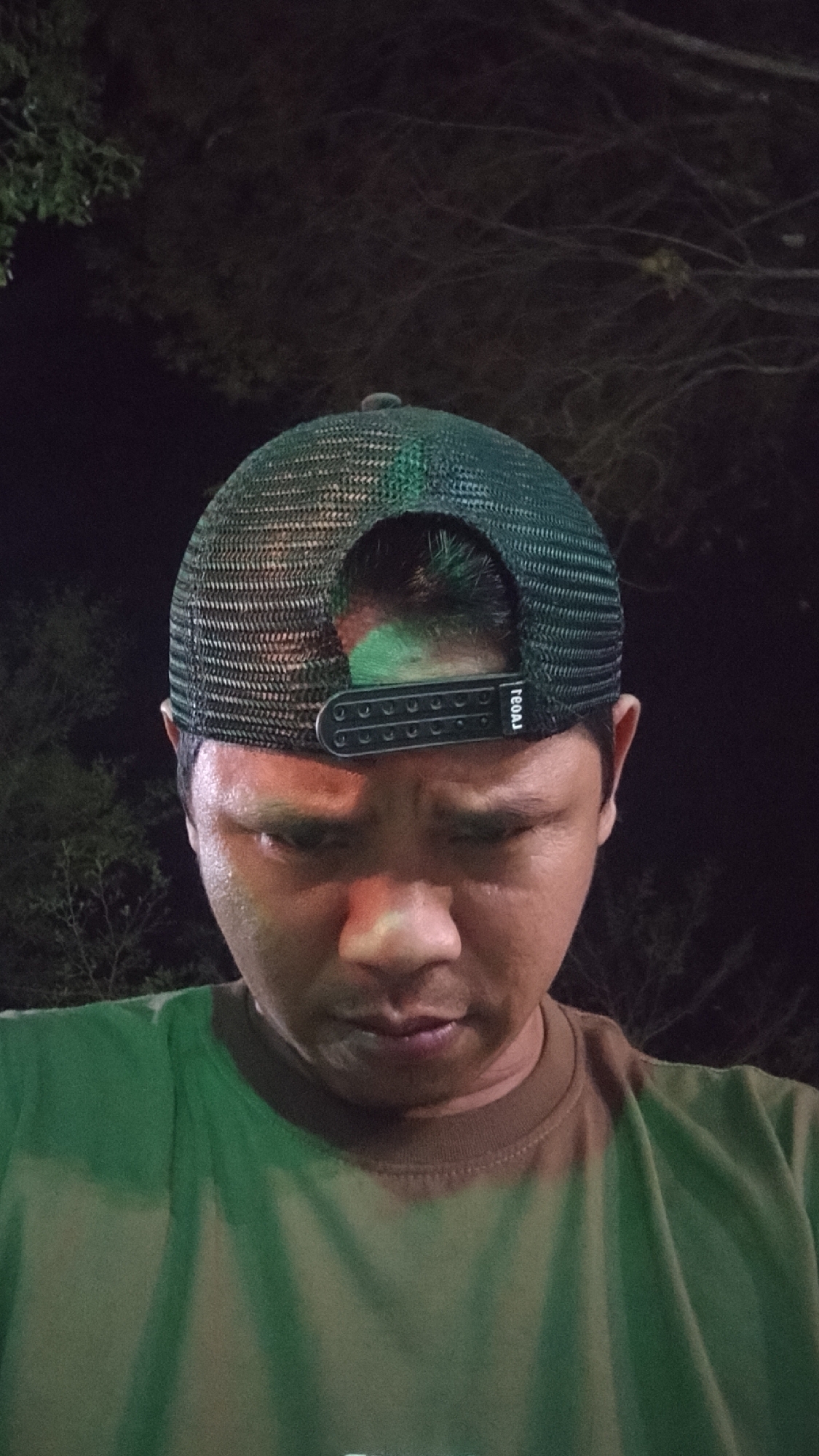 A man wearing a dark green mesh trucker hat and a green t-shirt is looking down in this close-up shot. His face is shiny with sweat. The background is dark and appears to be outdoors at night, with out-of-focus trees and branches visible. The man's head is tilted down, obscuring his eyes. A green reflective light is visible on the top of his head. The hat has a black band with a white logo on the back. The t-shirt has a brown collar and some lighter green discoloration on the front, possibly from sweat. The overall lighting is dim, with a green cast from the reflective light.