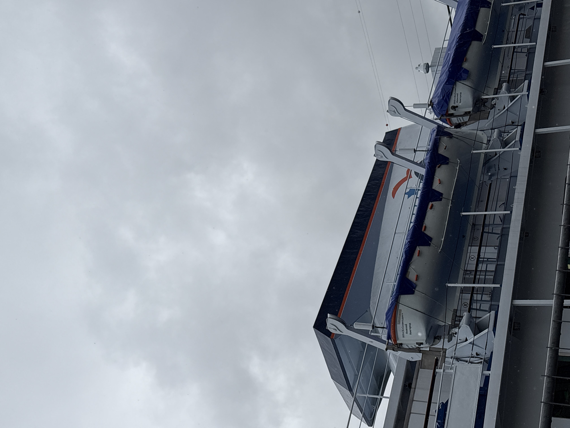The image shows two lifeboats hanging on the side of a ship against a cloudy sky. The lifeboats are white with blue covers. Text in Russian "000«СК»Созвездие» Ножкчий Новгород" is printed on the lifeboats. The ship's metal structures are visible. The location is likely in Russia, possibly the city of Saint Petersburg, given the Russian text on the lifeboats.