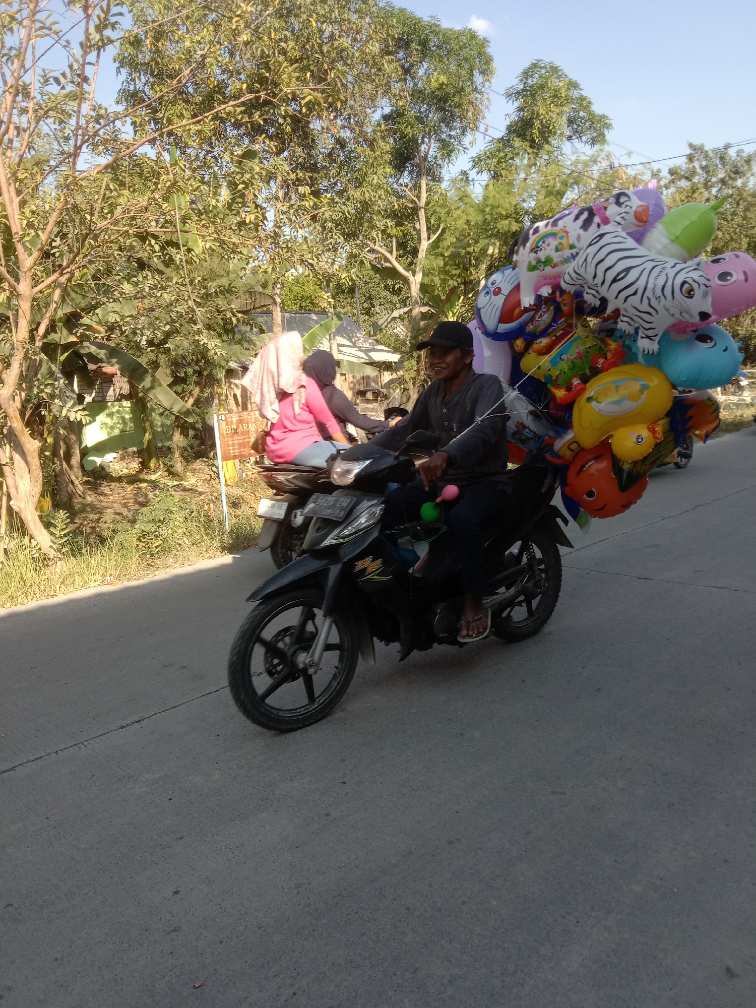 In Tulungrejo, Indonesia, a man wearing a black cap and a dark shirt rides a black motorcycle on a paved road. He is carrying a large bunch of colorful balloons on the back of the motorcycle. The balloons are shaped like various animals, including a tiger, a dog, and a duck, as well as cartoon characters. A woman wearing a pink shirt and a hijab is a passenger on the motorcycle. The background shows trees, vegetation, and buildings. The sky is clear and blue.