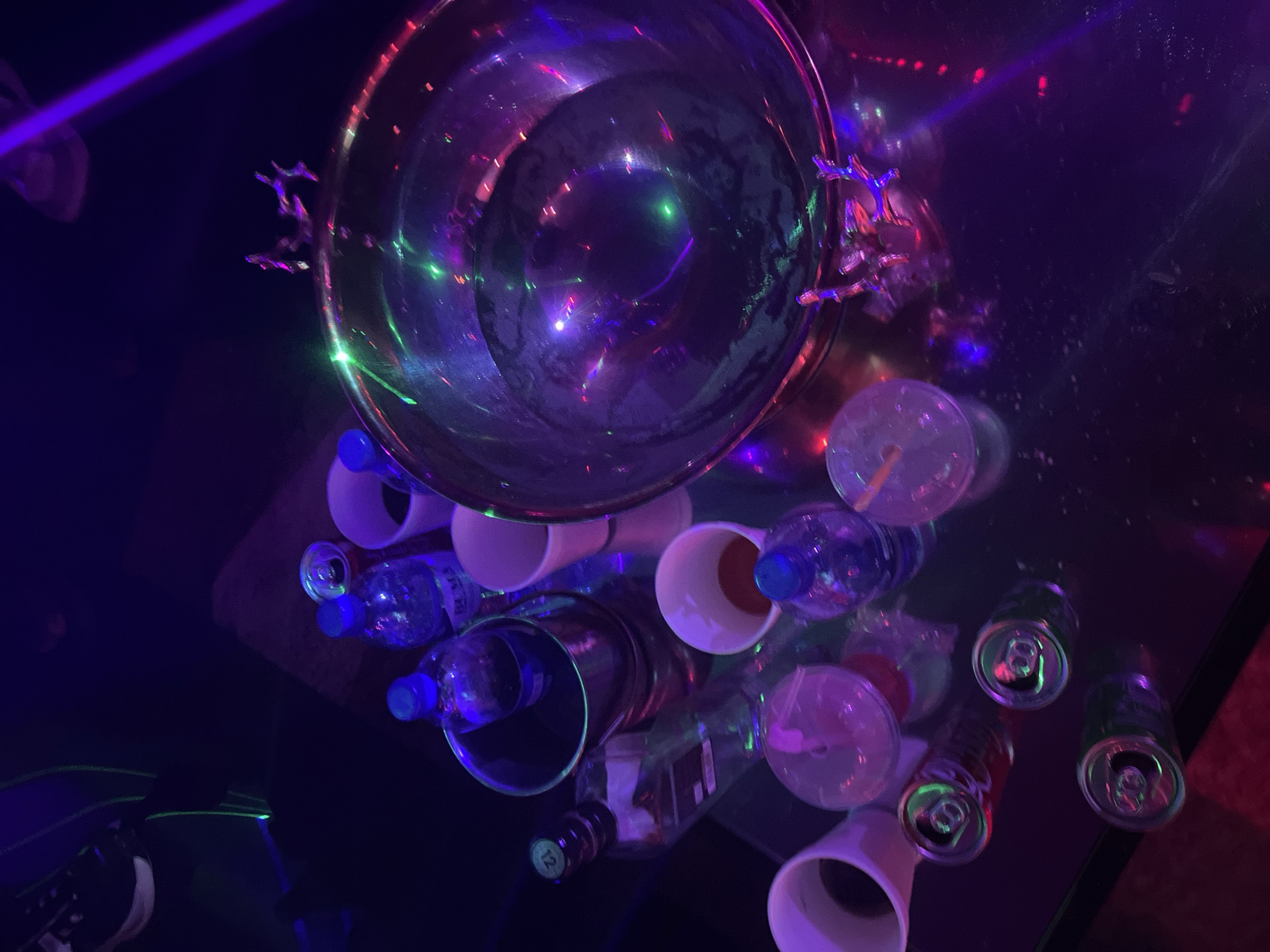 The image depicts a tabletop scene, likely in a bar or club in Jalingo, Nigeria, given the context. The table is covered in a reflective surface, strewn with discarded beverage containers. A large metal punch bowl is positioned at the top, reflecting purple and green lights. Discarded water bottles, plastic cups, and soda cans are scattered around it. A glass bottle with a dark label is partially visible. A small section of a person's shoe is visible in the bottom left corner. The overall lighting is dark, with neon-colored highlights from lighting effects.