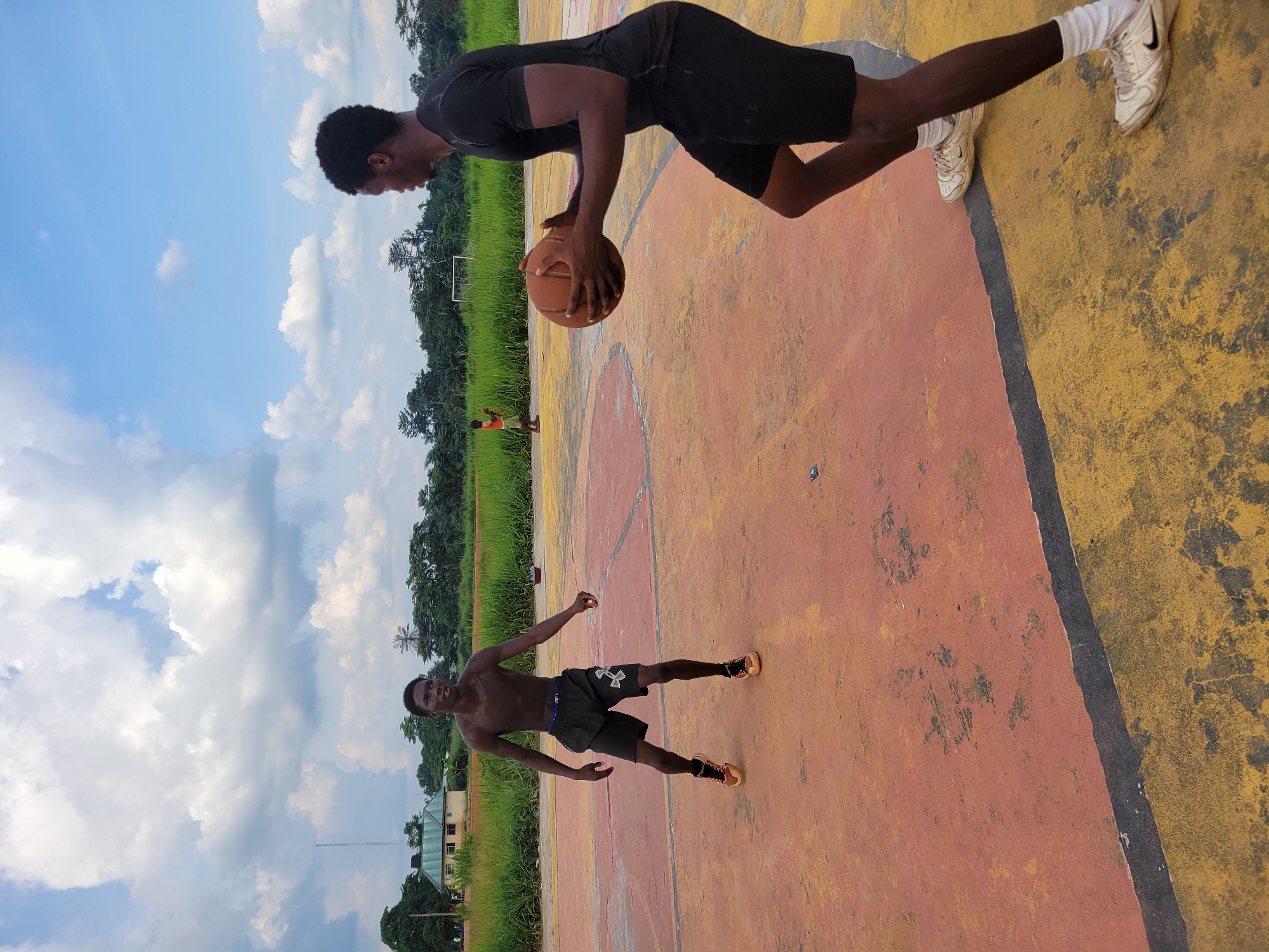 The image depicts two young men playing basketball on an outdoor court in Egbagu, Nigeria. The court has a faded yellow and orange surface with black lines. In the foreground, one man is in a push-up position, his body angled parallel to the court. He is shirtless and wearing dark shorts and orange sneakers. His gaze is directed towards the viewer.
In the background, another man, also shirtless, is leaning over to catch a basketball. He is wearing black shorts and white socks with sneakers. The basketball is a prominent orange sphere held in his hands.
Beyond the court, a line of lush green grass and trees stretches across the landscape. A white soccer goal can be seen in the distance, suggesting a multi-purpose sports area. The sky is partly cloudy with dramatic white cumulus clouds against a vibrant blue backdrop. The lighting suggests it is daytime, likely late morning or early afternoon, with the sun casting distinct shadows. The overall atmosphere appears to be one of casual recreation and physical activity in a tropical environment.