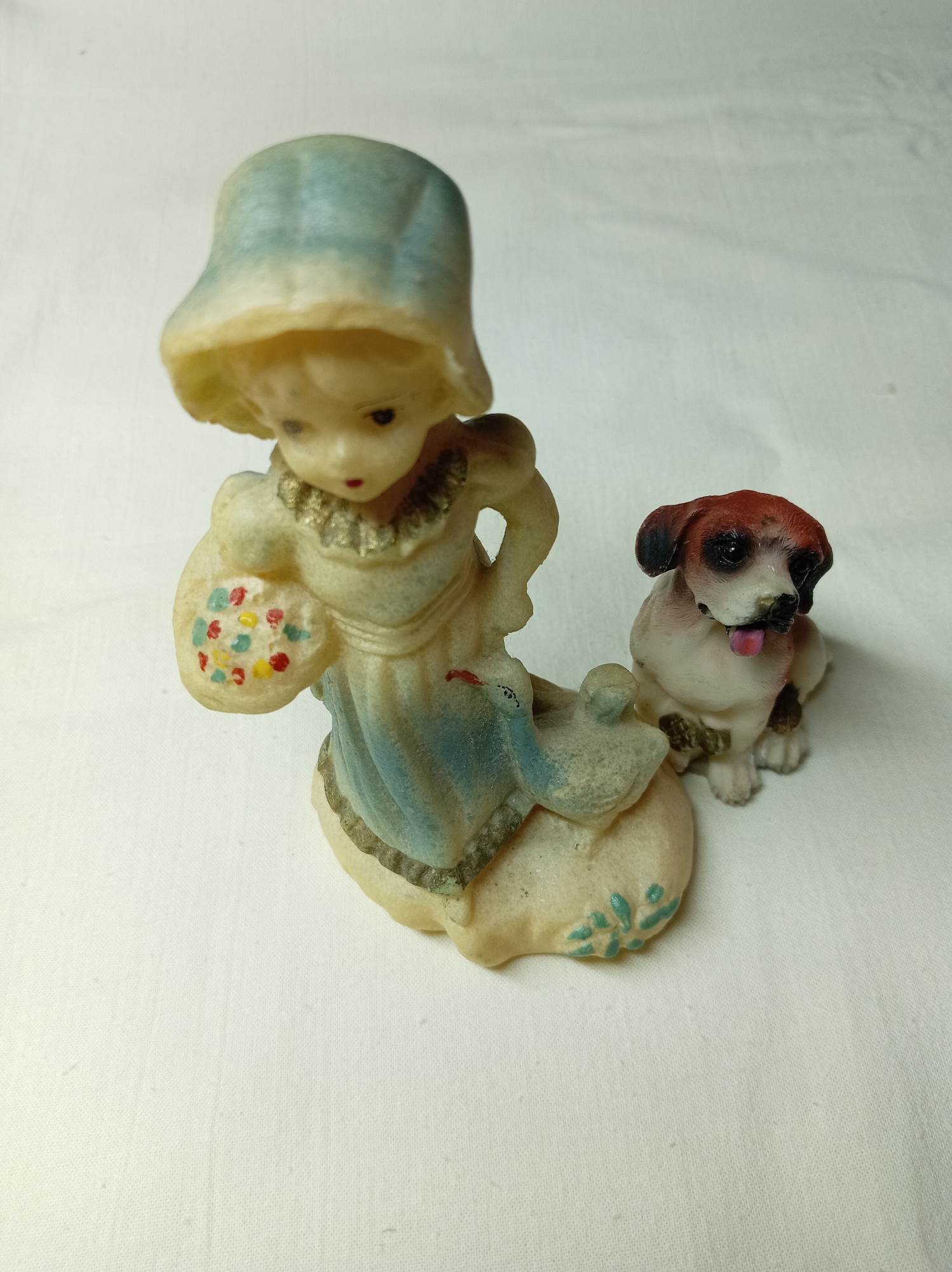 A figurine of a girl in a blue hat and dress, holding a basket of flowers, is positioned next to a small dog figurine. The figurines are placed on a white fabric surface. There is no indication of the location in the city of Bugulma, Russia.