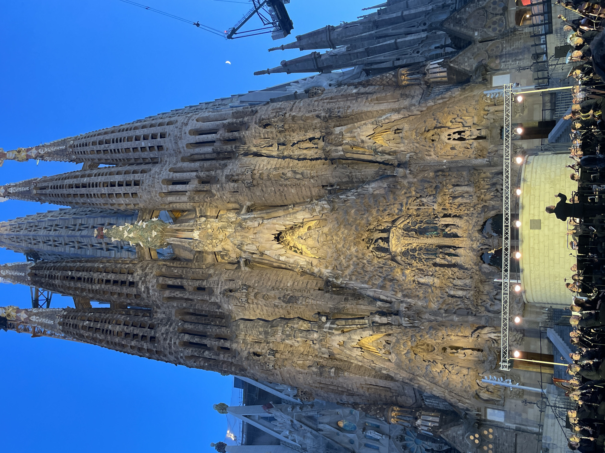The image captures a wide-angle, low-light exterior shot of the Sagrada Familia in Barcelona, Spain, at dusk. The iconic basilica's intricate facade, illuminated from below, dominates the frame. Construction cranes are visible in the upper right portion of the image, indicating ongoing work on the structure. In the foreground, a large orchestra is set up on a stage directly in front of the basilica, with a conductor standing and gesturing to the musicians. The orchestra members are dressed in black attire and are seated with their instruments, appearing to be in the midst of a performance. The sky is a deep twilight blue with a sliver of a crescent moon visible. The overall scene depicts a cultural event taking place at a significant architectural landmark.