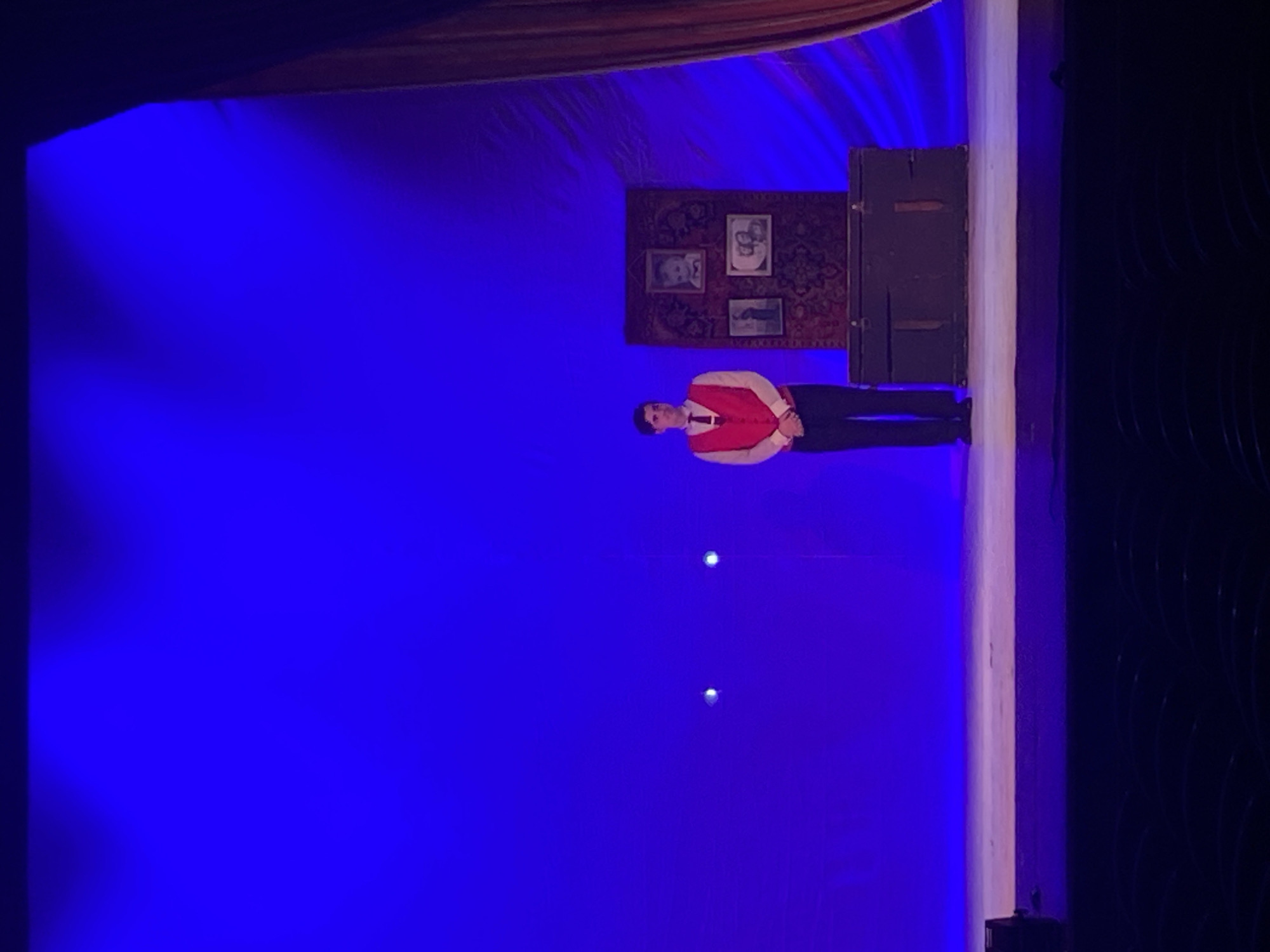A man in a red vest and black pants is standing on a stage with a blue backdrop. Behind him is a wall with a rug and three framed pictures. To the right of the man is a dark brown cabinet. The stage is set inside a building. Rusanovo, Russia is not specified in the image.