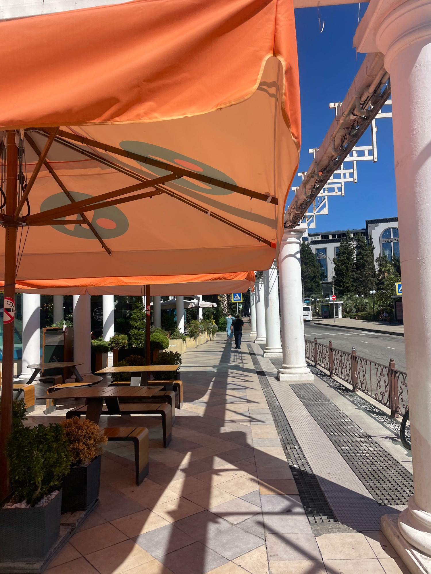 An outdoor seating area with orange umbrellas is depicted in Sochi, Russia. The scene is a sunny day, with shadows cast by the umbrellas and columns onto a patterned tiled walkway. Several wooden tables and benches are arranged under the umbrellas. Beyond the walkway, a street is visible with traffic, a bus stop, and buildings. Two people are walking on the sidewalk in the distance. The area appears to be a commercial or tourist district, possibly a cafe or restaurant.