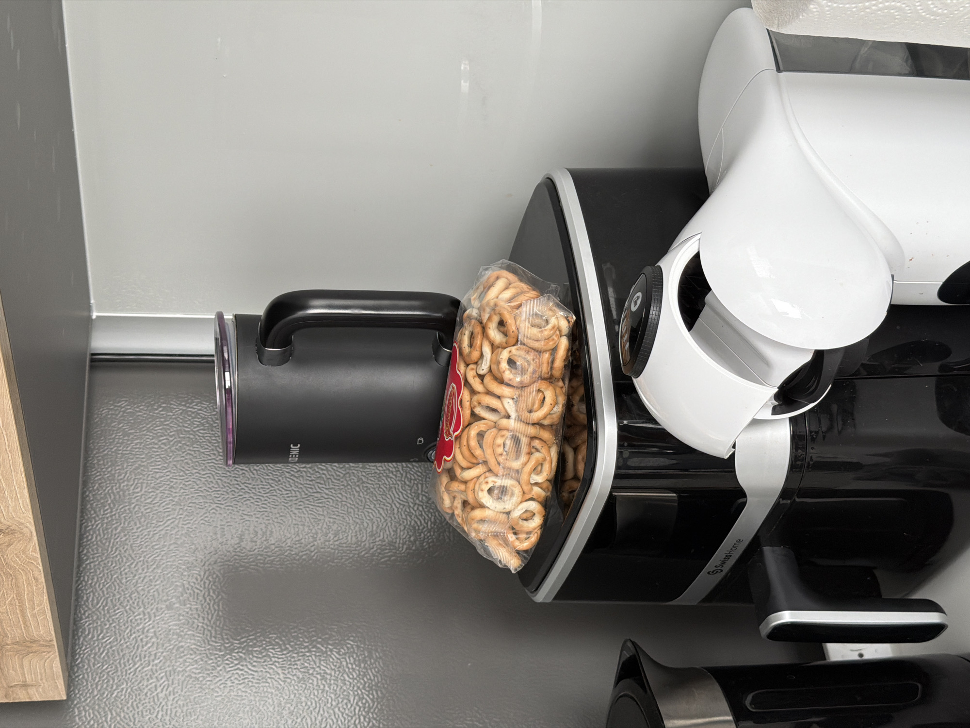 A close-up, eye-level, indoor shot shows a collection of kitchen appliances and a bag of pretzels. On the left, a black electric kettle with a silver lid and handle is partially visible. In the center, a black coffee maker with a silver trim and handle is stacked with a black travel mug on top. A clear plastic bag filled with small, ring-shaped pretzels is placed on top of the coffee maker. To the right of the coffee maker is a white coffee pod machine. The background consists of a gray, textured wall and a light wood shelf above. The scene is brightly lit, with no people present. The image does not provide any information about its location in Finestrat, Spain, as it is a generic kitchen setting.