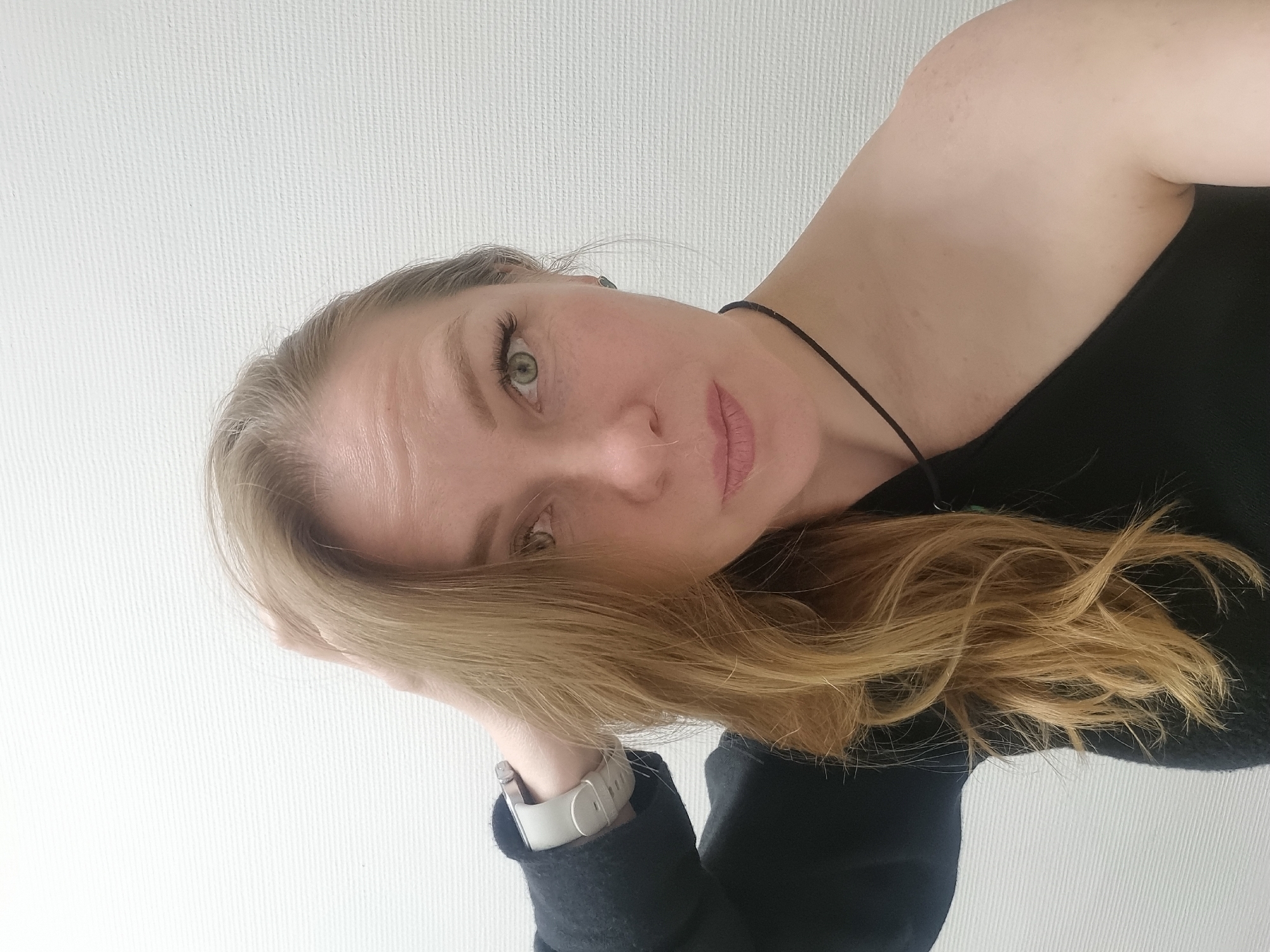 A woman with light blonde hair and green eyes is pictured in a selfie-style image. Her head is tilted to the right. Her left hand, wearing a white smartwatch, is positioned near her face, supporting it. She is wearing a black top. A thin black necklace is visible. The background is a plain white wall. The location is unknown. There is no evidence indicating it is in Kemul, Russia.