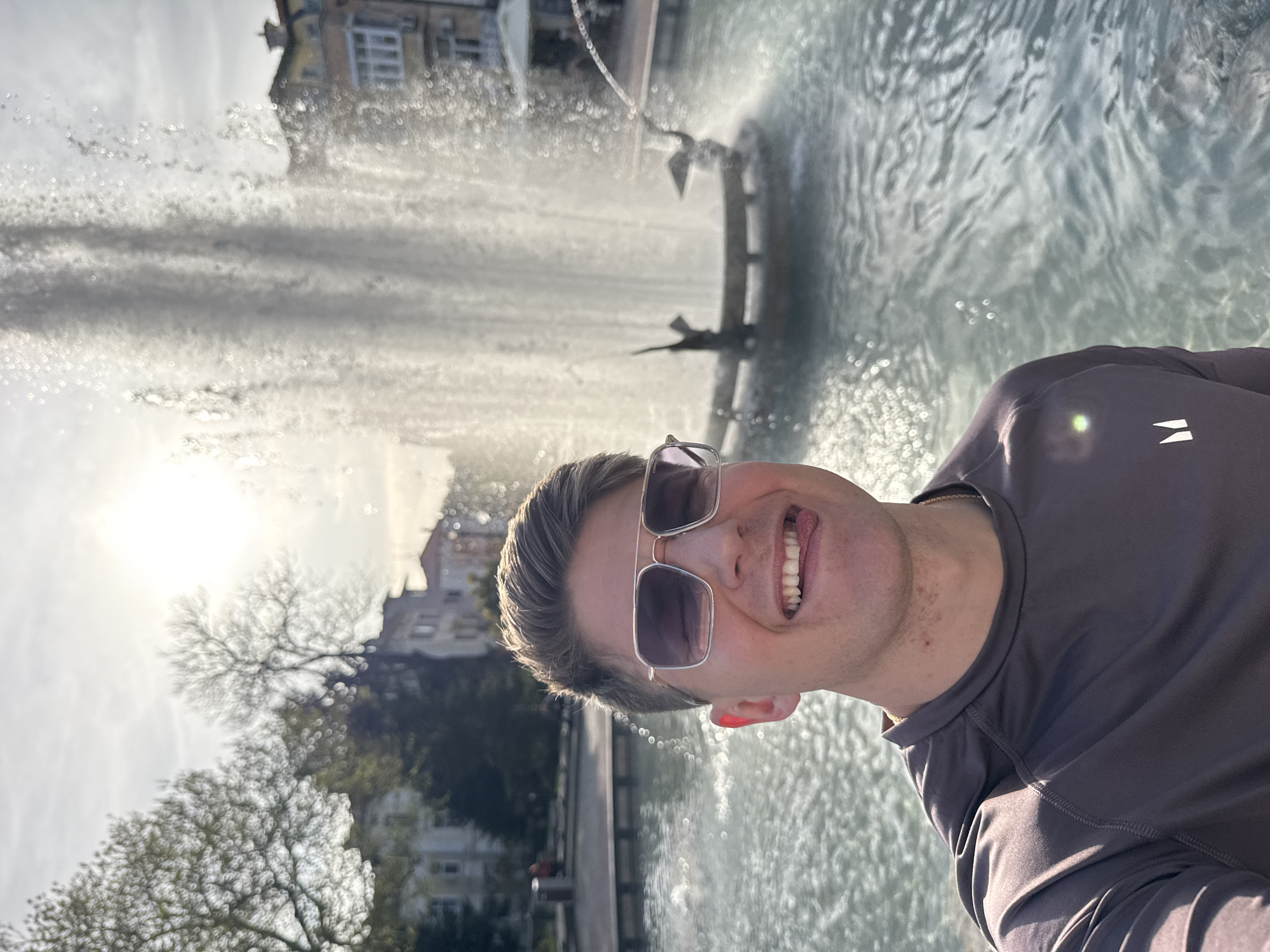 A man is taking a selfie in front of the Plovdiv Singing Fountains in Bulgaria. The fountain is active, spraying water high into the air. The man is wearing sunglasses and a gray shirt and is smiling with his tongue slightly out. The background includes trees, buildings, and the sun.