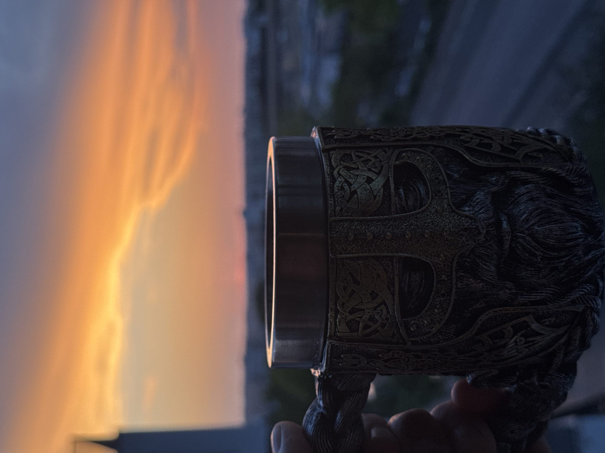 A close-up shot features a hand holding a Viking-themed mug against a sunset sky. The mug is intricately designed with a helmeted face and braided handle, rendered in a metallic, aged finish. The background displays a blurred cityscape with buildings and a road, bathed in the warm, gradient hues of the setting sun, transitioning from orange to purple.