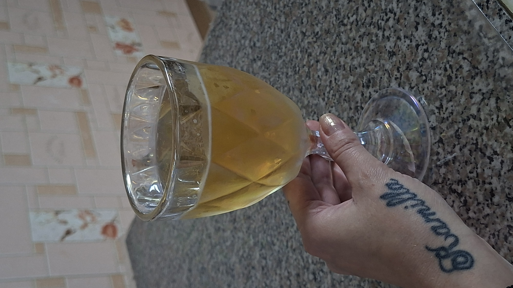 A hand with a tattoo of the name "Kamilla" on the wrist is holding a clear glass filled with a pale yellow liquid. The glass has a diamond-patterned texture. The hand has nails painted with gold-colored polish. The glass and hand are resting on a speckled gray countertop. In the background, a tiled wall with faint floral or patterned designs is visible. The scene appears to be indoors, possibly in a kitchen or dining area. There is no visible indication of the location within the city of Myadzyel, Belarus.