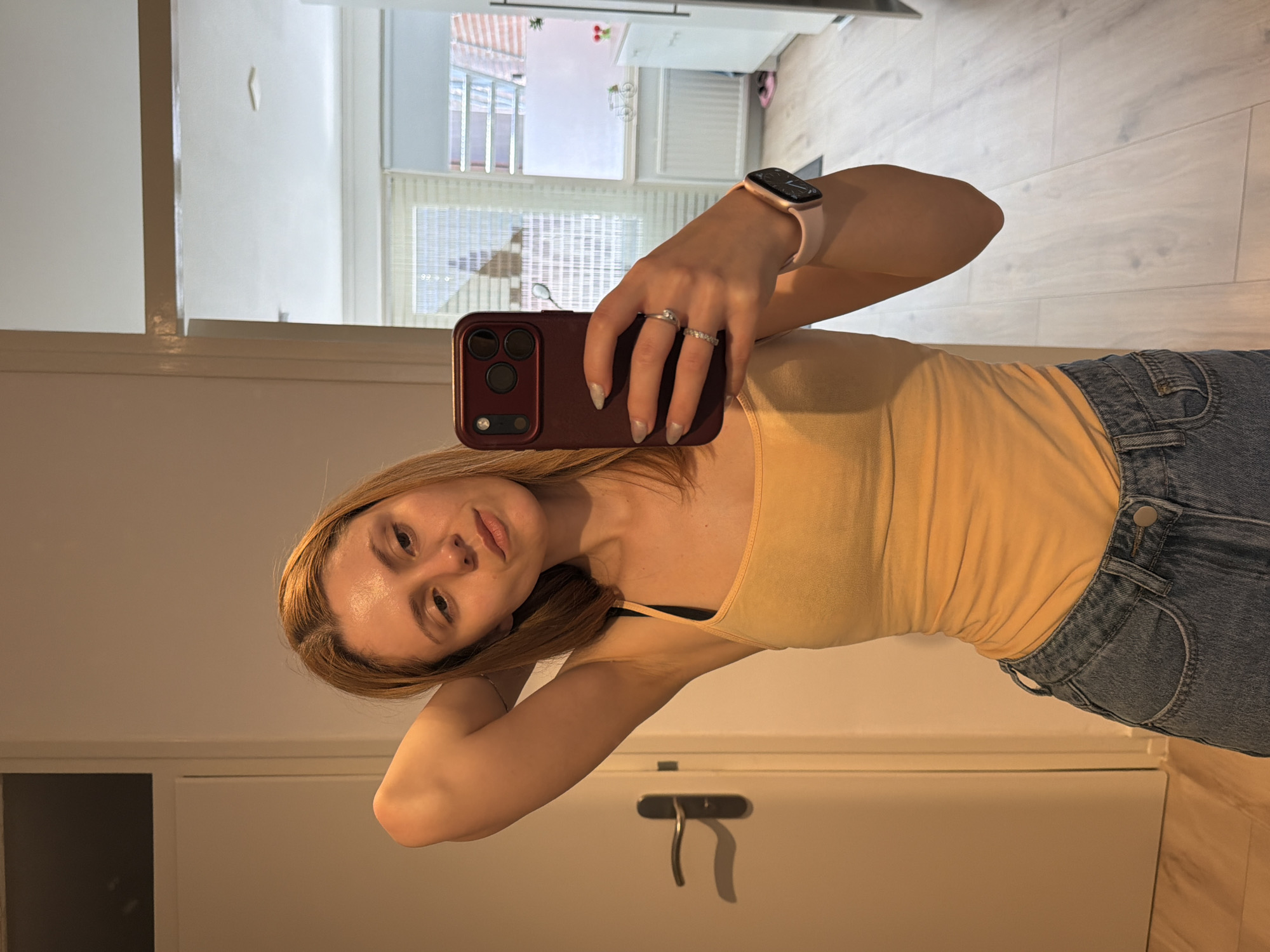 A young woman with blonde hair, wearing a yellow tank top and blue jeans, is taking a mirror selfie. She is standing in what appears to be a hallway or entryway. Her right arm is raised, with her hand behind her head, and she is looking at her reflection in the mirror. On her left wrist, she is wearing a pink smartwatch. She has a ring on her left ring finger. In the background, a door with a silver handle is visible to her left. To her right, through a doorway, a room with white cabinets and a window with blinds can be seen. The floor is made of light-colored wood. The overall lighting suggests it is daytime. The location is not specified in the image.