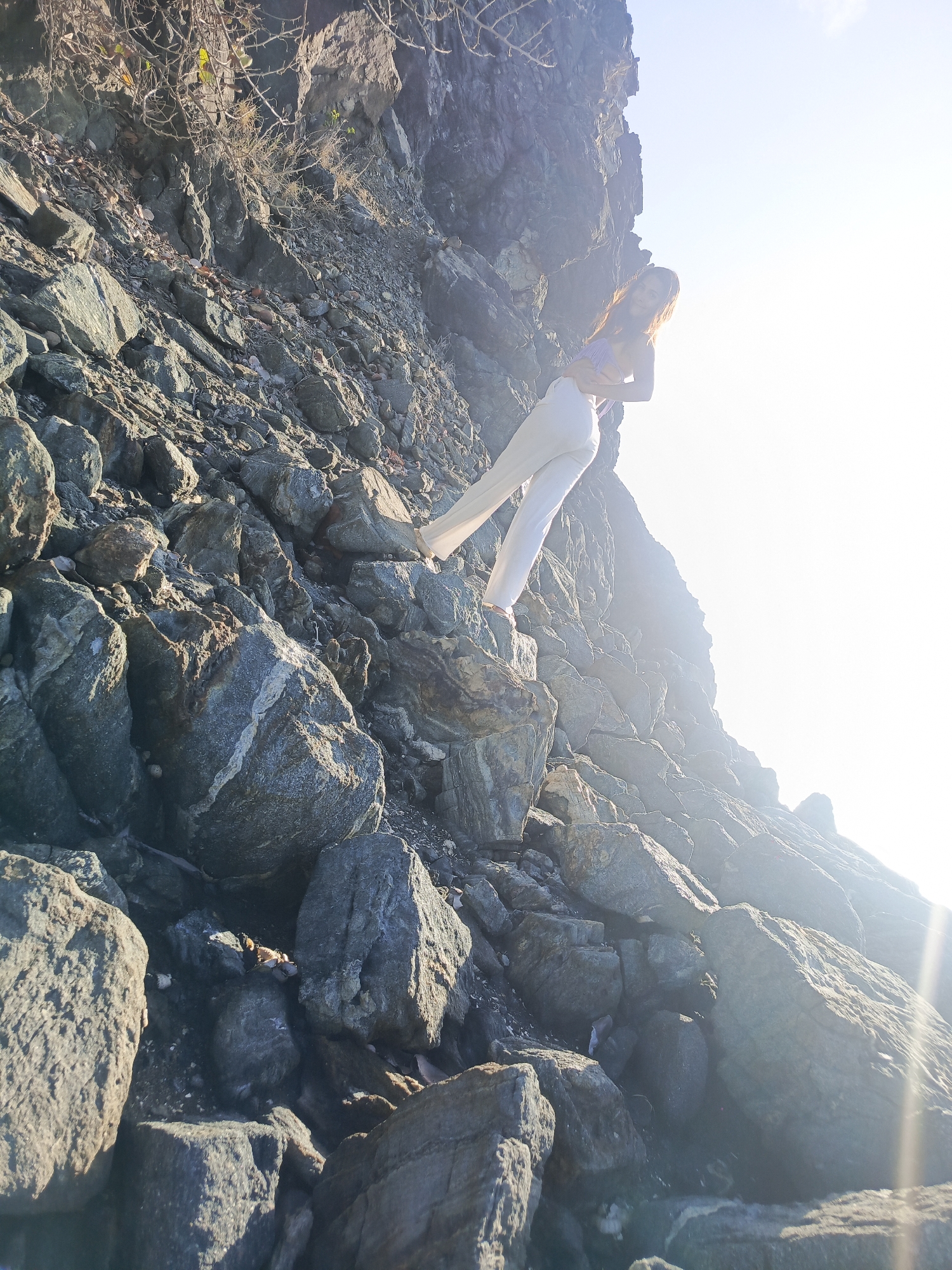 A young woman is climbing up a rocky cliff. She is wearing a purple top and white pants. The sun is shining brightly, creating a lens flare in the image. The cliff is made up of large gray rocks. There are some dry branches and leaves on the cliff. The location is a natural outdoor setting.